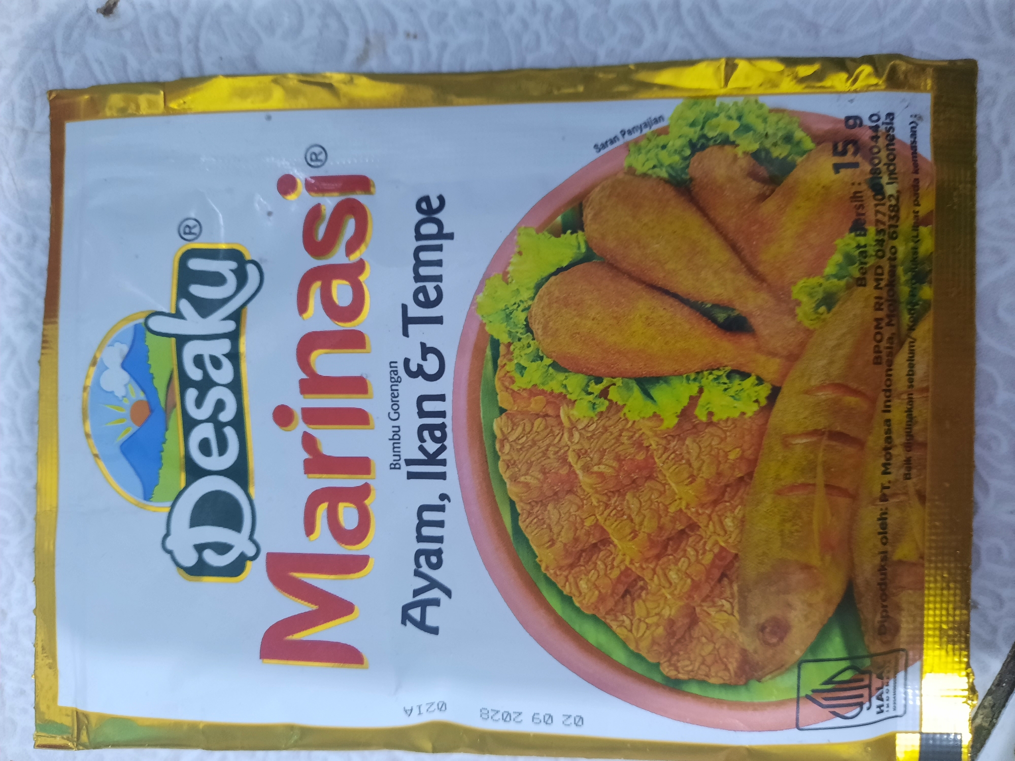 The image shows a sealed sachet of "Desaku Marinasi" seasoning. The sachet is gold and white with a colorful illustration of fried chicken, fish, and tempeh. Text on the sachet indicates it is for marinating chicken, fish, and tempeh and includes production details, net weight (15g), and a Halal certification logo. There is no indication of the city of Lhokseumawe, Indonesia, in the image.