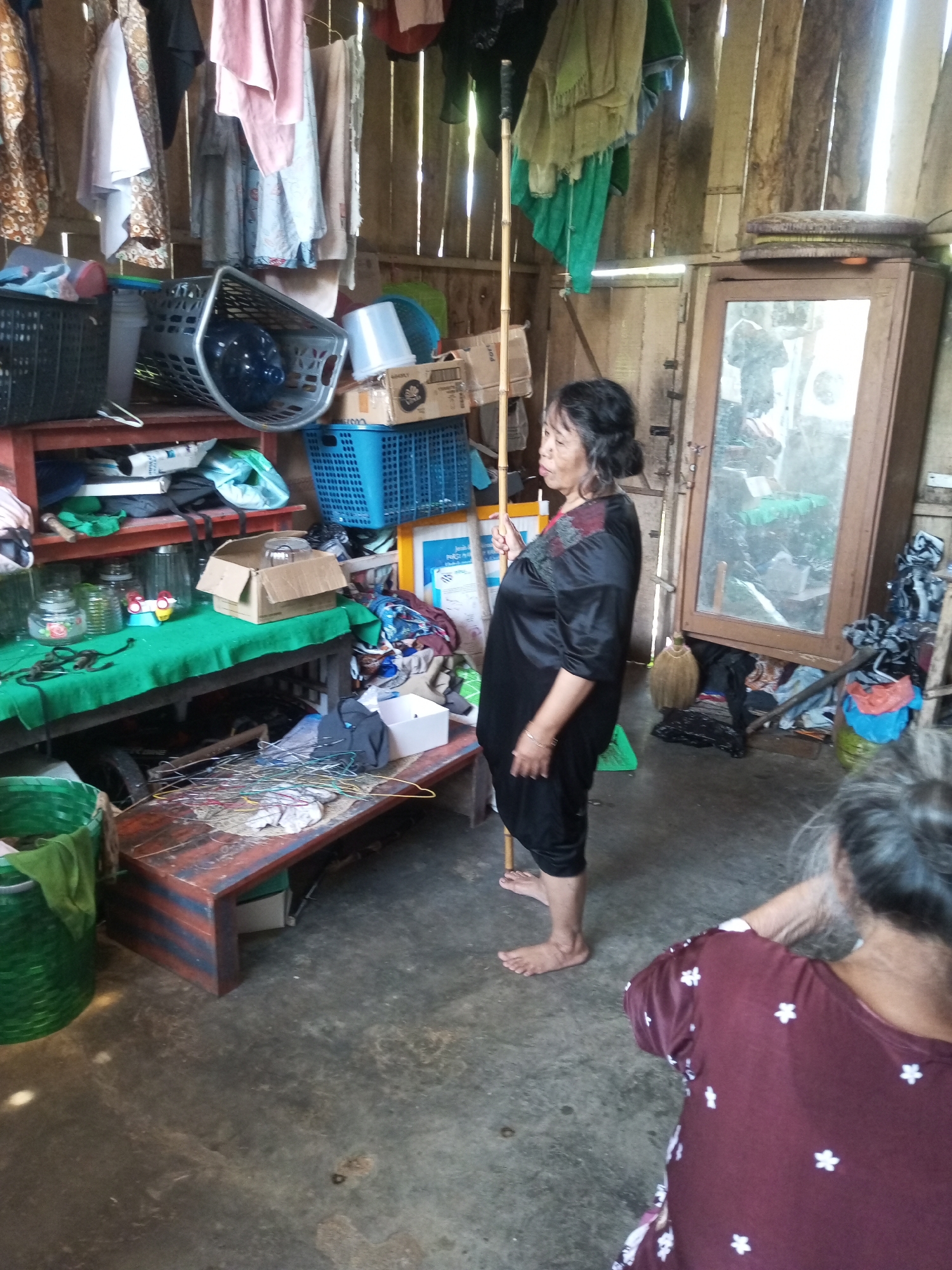 In Krasak, Indonesia, within a rustic interior space constructed with vertical wooden planks, a woman in a black satin outfit stands holding a long bamboo pole. Her hair is dark and pulled back, and she wears subtle jewelry. In the foreground, another woman with grey hair is visible from behind, wearing a maroon shirt adorned with white floral patterns. The room is cluttered with various items, including laundry hanging from overhead structures, shelves filled with baskets and containers, and a table with a green cloth on it displaying jars and a cardboard box. A large wooden cabinet with a mirror is positioned against the back wall. The floor is a rough concrete surface.