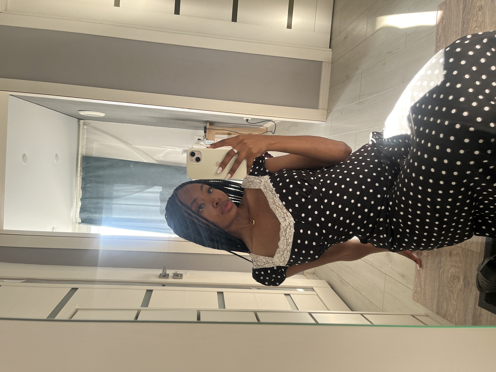 A young Black woman with braided hair takes a mirror selfie in an indoor setting. She is wearing a black and white polka dot dress with a lace trim. The woman is seated, and her legs are crossed. She holds a white iPhone in her right hand. The background consists of a light-colored wall, a door, and a window with blue curtains. The floor is made of light-colored wood planks. Sunlight streams in from the window, casting a bright rectangle on the floor.