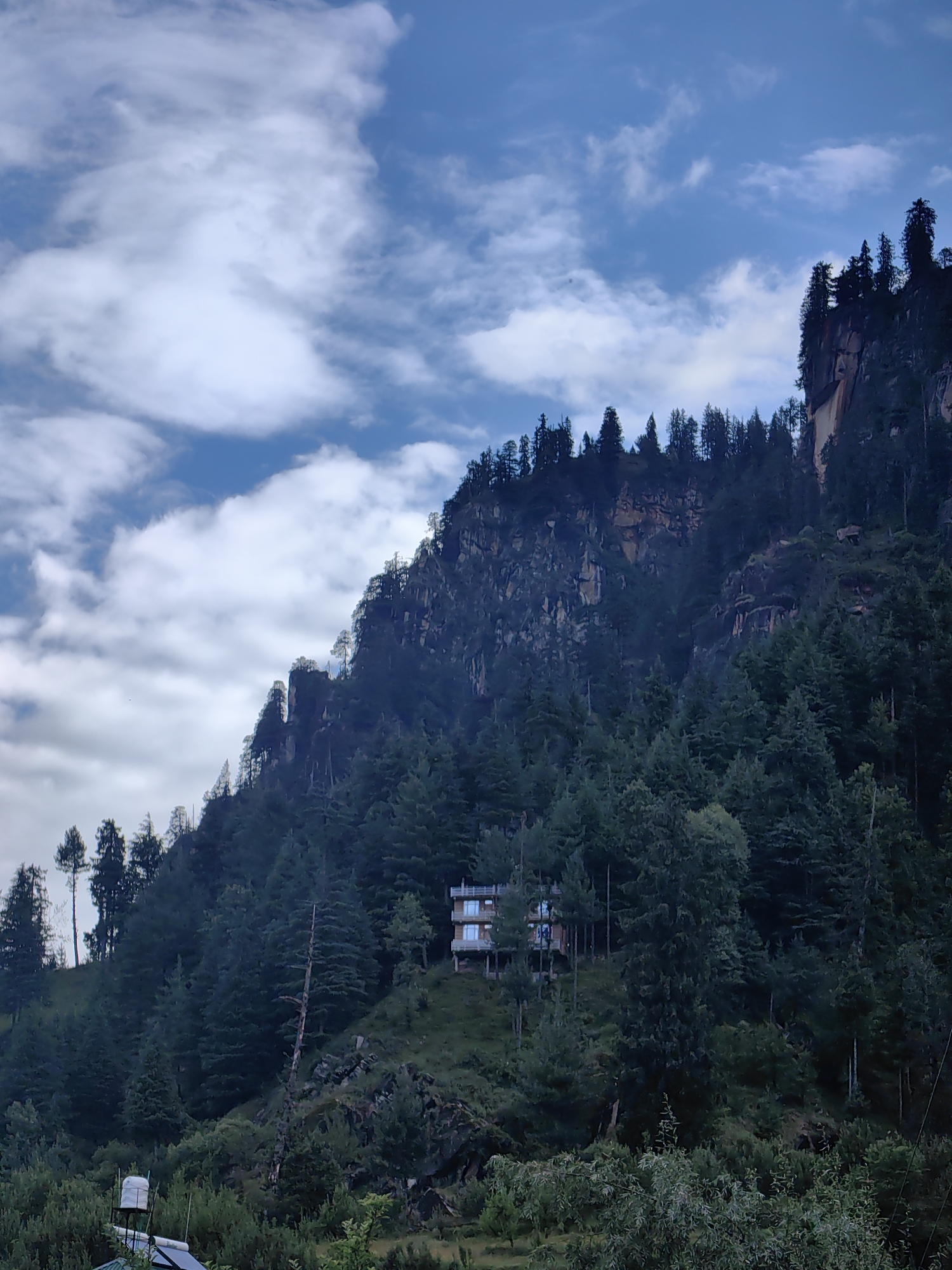A multi-story building with a light-colored facade and multiple balconies is situated on a hillside covered in dense coniferous trees. The building is surrounded by lush greenery and appears to be built into the slope of the mountain. In the foreground, there are more trees and some smaller structures, including what appears to be a water tank on a platform. The background features a steep, rocky mountain face with more trees growing on its slopes. The sky above is blue with scattered white clouds. The scene depicts a mountainous landscape with a human dwelling integrated into the natural environment, likely in a village or town setting in Vashisht, India, given the context provided.