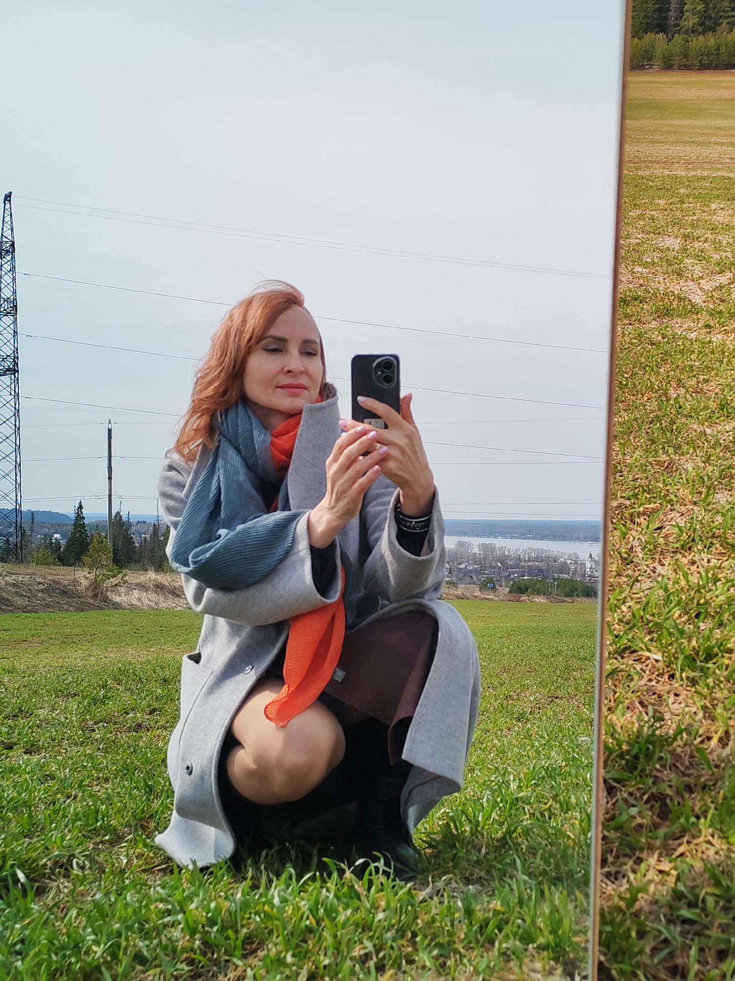A woman with red hair is squatting in a grassy field, holding a smartphone to take a selfie in a mirror propped up beside her. She's wearing a grey coat with an orange and blue scarf. An electricity pylon is visible in the background under a blue sky. In the mirror reflection, a field and trees are visible as well as a body of water and a city with churches. This location appears to be in Nechkino, Russia.