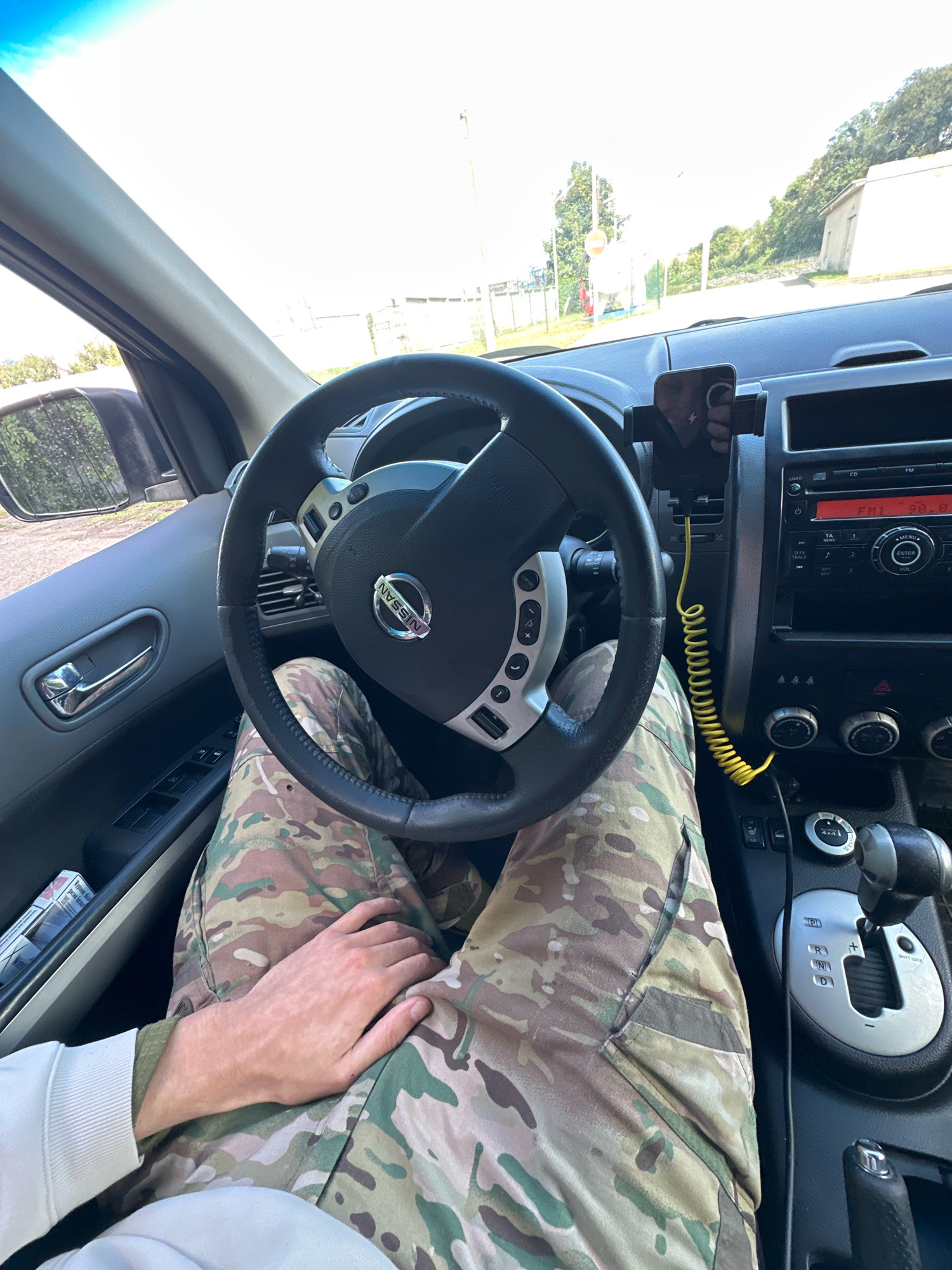 The image shows the interior of a car, likely a Nissan, seen from the driver's perspective. The driver's hands are visible resting on their lap, which is covered by camouflage pants. The steering wheel, dashboard, and center console are prominent features. A smartphone is mounted on a holder in front of the dashboard, connected to a yellow charging cable. The car's radio is on, displaying "FM1 90.8". The gear shift is visible, indicating the car is in park. The exterior environment suggests an outdoor setting, possibly near a rural or industrial area with buildings and trees visible through the windshield. The sky is bright and overcast. The location "Shatava, Ukraine" is not explicitly indicated within the image itself.