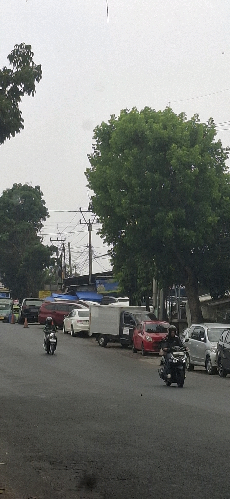 In Rancagoong, Indonesia, a street scene unfolds under an overcast sky. The asphalt road shows signs of wear, with patches and cracks visible. Several vehicles are parked along the right side of the road, including a silver SUV, a black car, and a red hatchback. A white box truck is also present, positioned behind the red hatchback. To the left, a white sedan is parked, followed by a maroon SUV and a black SUV. Two traffic cones are placed on the road.
Two individuals are riding motorcycles. The first, on the left, is on a black scooter, wearing a helmet and dark clothing. The second, on the right, is on a black motorcycle, also wearing a helmet and dark clothing.
Lush green trees line the side of the road, their leaves dense and full. Utility poles with numerous wires are visible in the background, suggesting an urban or semi-urban environment. In the distance, there are various structures, some appearing to be shops or stalls, covered with blue tarpaulins. The overall atmosphere is one of a typical day in a busy Indonesian town.