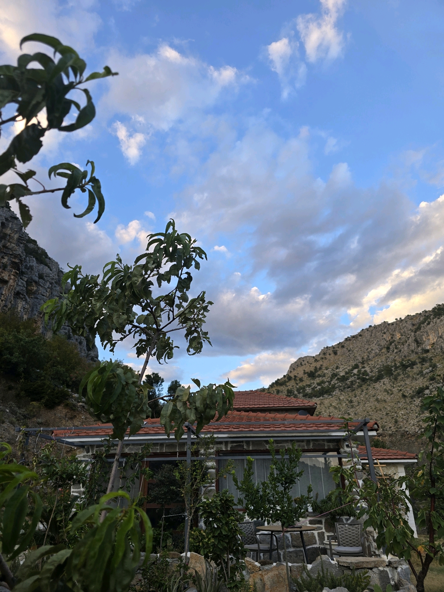 The image depicts a two-story house with a red tile roof, set against a mountainous backdrop under a partly cloudy sky. The house is constructed with stone walls and has large windows. In the foreground, there are trees with green leaves, some of which are out of focus. A small seating area with a table and two chairs is visible on a stone patio in front of the house. The surrounding environment appears to be a natural, somewhat rugged landscape with rocky slopes and sparse vegetation. The scene is captured during the daytime, with soft lighting suggesting either early morning or late afternoon. The location is described as Podgorica, Montenegro.