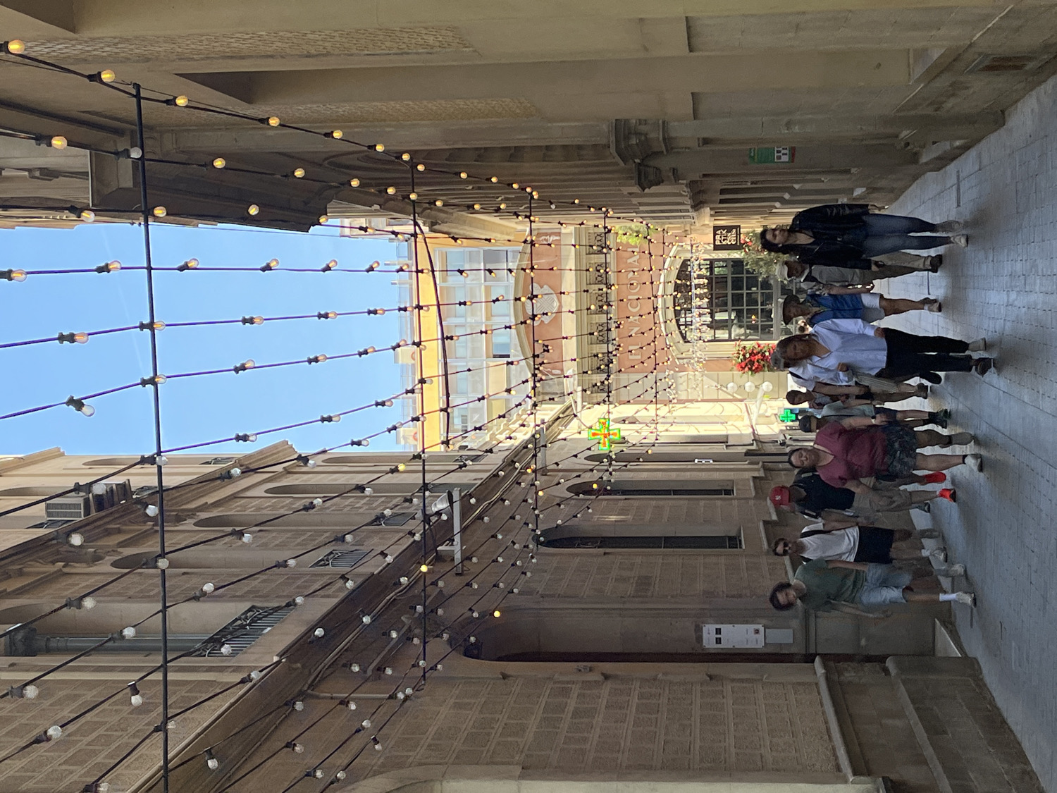 The image shows a narrow pedestrian street in Barcelona, Spain, bathed in bright daylight under a clear blue sky. Strings of decorative, bulb-style lights are strung overhead, stretching across the street between the buildings, creating a canopy effect.
A group of approximately ten people are visible on the right side of the street, walking away from the viewer. They appear to be casually strolling, with varied attire suggesting a warm, sunny day. The buildings lining the street are multi-story, featuring light-colored stone or stucco facades with windows and balconies.
Notable details include the bright, sunny weather and the characteristic architecture of a European city. On the left side of the street, a building clearly displays the word "FARMACIA" (Spanish for pharmacy) and a prominent green cross sign, a common symbol for pharmacies in many European countries. Another, less clear sign appears to read "LESLIES" or similar text near the pharmacy. The street itself is paved with light-colored flagstones. The overall scene depicts a lively, yet relaxed, urban environment.