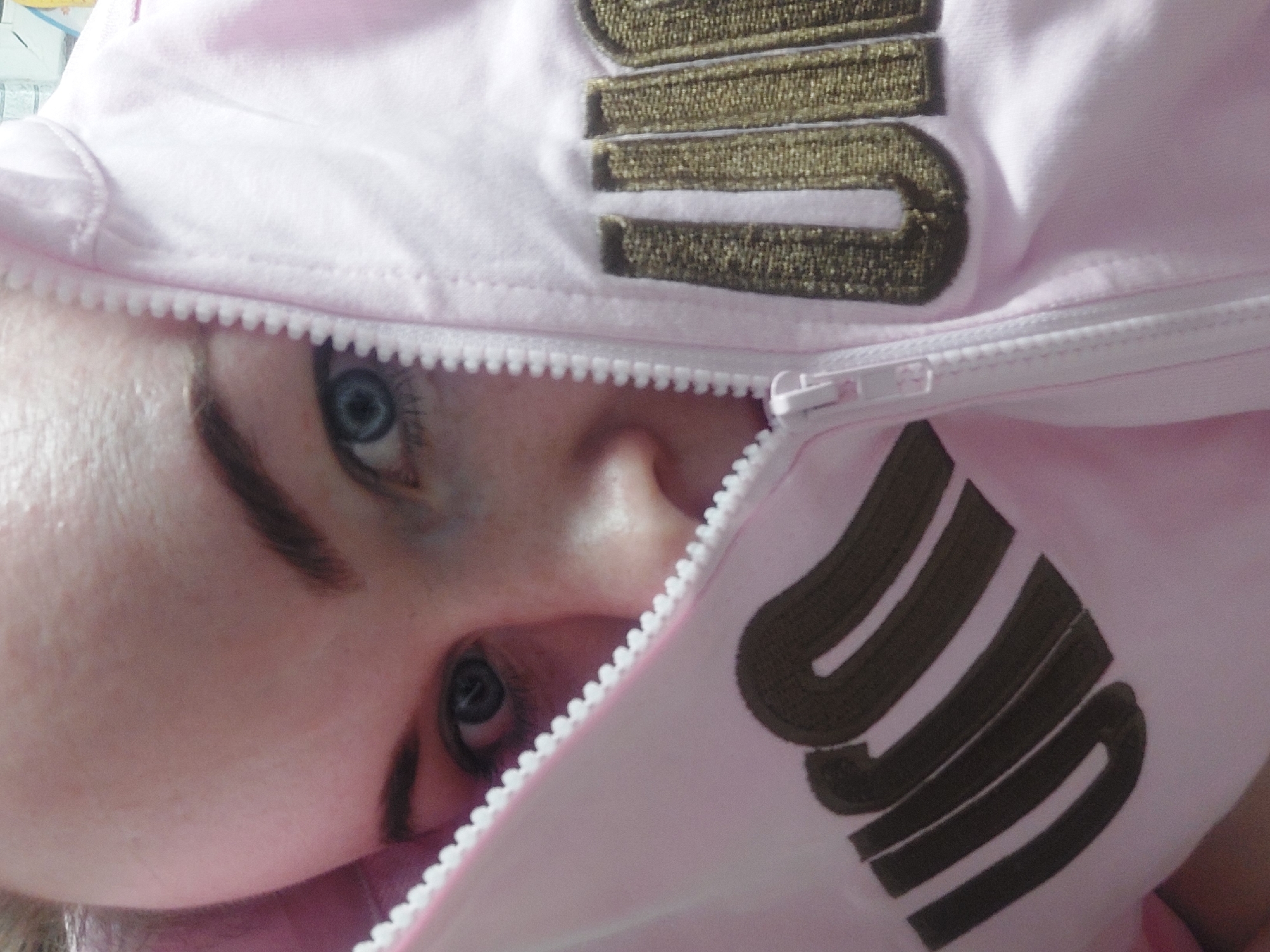 A close-up, slightly angled image focuses on a person's face, partially obscured by a pink hooded jacket. The jacket's hood is partially zipped, framing the person's face. The jacket is embroidered with brown lettering. The person has fair skin, defined eyebrows, and blue eyes, which are the most visible feature. The person's nose and part of their cheek are also visible. The overall composition is intimate and centered on the person's facial features. The location is indiscernible.