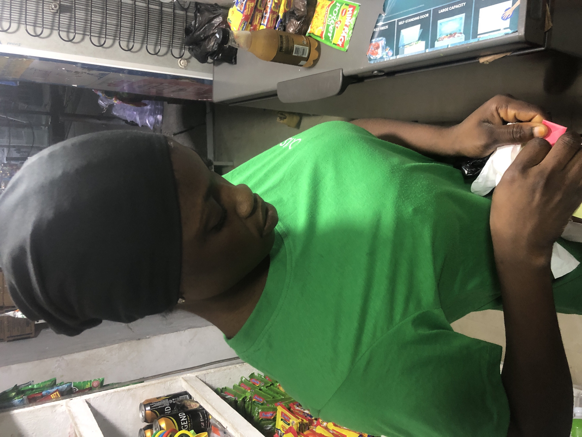 In Ado, Nigeria, a young Black woman wearing a green t-shirt and a black headwrap stands behind a counter. She is looking down at her hands, which are holding a small pink object and a white piece of paper. The counter is cluttered with various items, including a bottle of beverage, stacks of snacks, and a black trash bag. To the left of the woman, shelves are stocked with canned drinks and packaged goods. In the background, a refrigerator with a wire rack on its side is visible. The environment appears to be a small shop or convenience store.