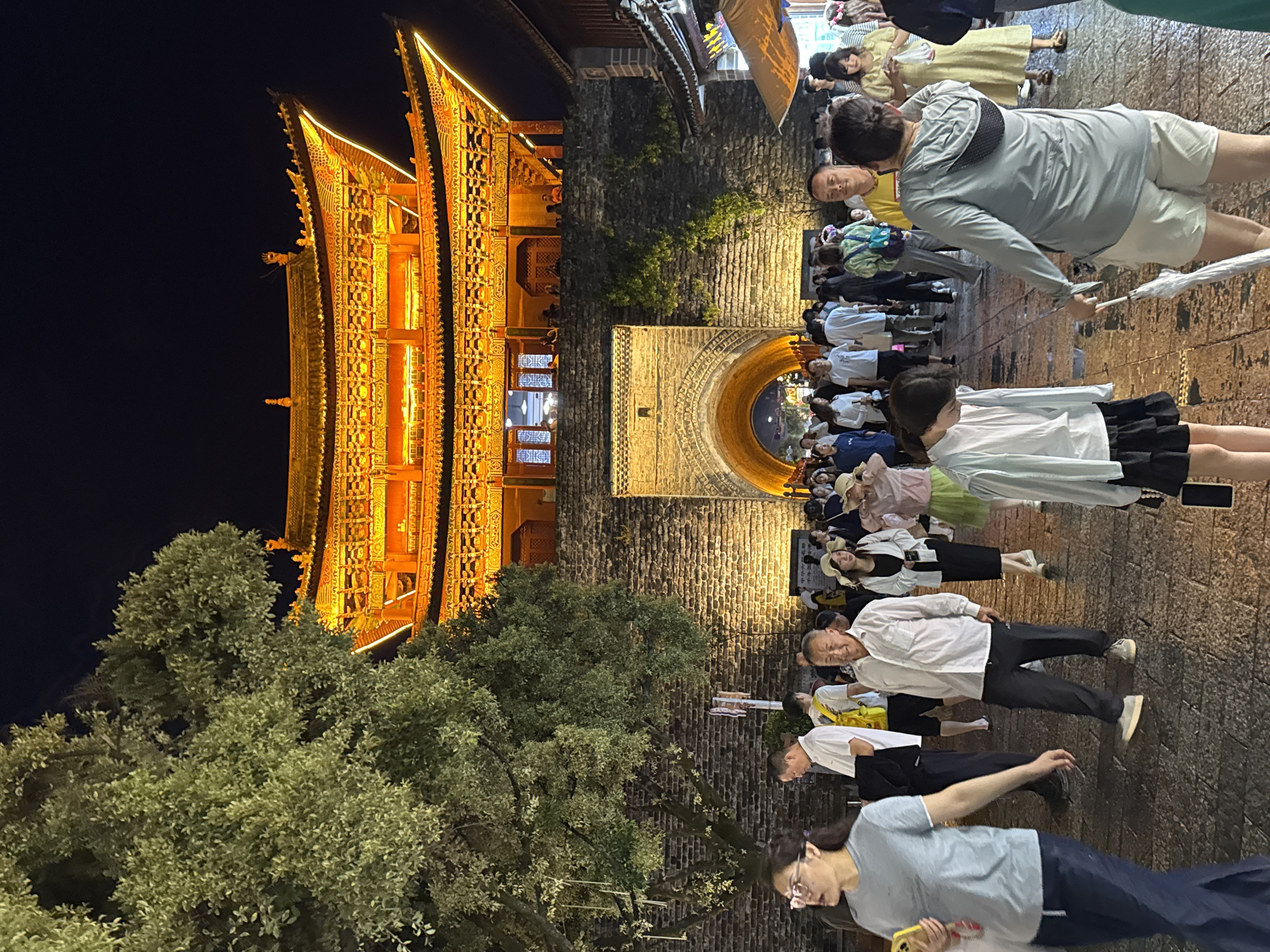 This image depicts the night scene at the ancient city gate of Dali, Yunnan Province, China. The gate, illuminated by warm yellow lights, features traditional Chinese architectural elements with layered, upturned eaves. A large brick wall surrounds the archway, which is also lit, creating a warm glow that spills onto the cobblestone ground. A lush green tree with dense foliage stands to the left, partially obscuring the gate and the night sky.
A diverse crowd of people is present, milling around and walking through the archway. They are dressed in casual summer clothing. Several individuals are in focus in the foreground, including a woman wearing glasses and a grey t-shirt, a man in a white shirt and black pants, and a woman in a light blue shirt and black skirt. Other people are seen further back, some holding umbrellas. The overall atmosphere appears to be lively and bustling, characteristic of a popular tourist destination at night. The ground is paved with cobblestones, and the lighting creates a sense of warmth and festivity.