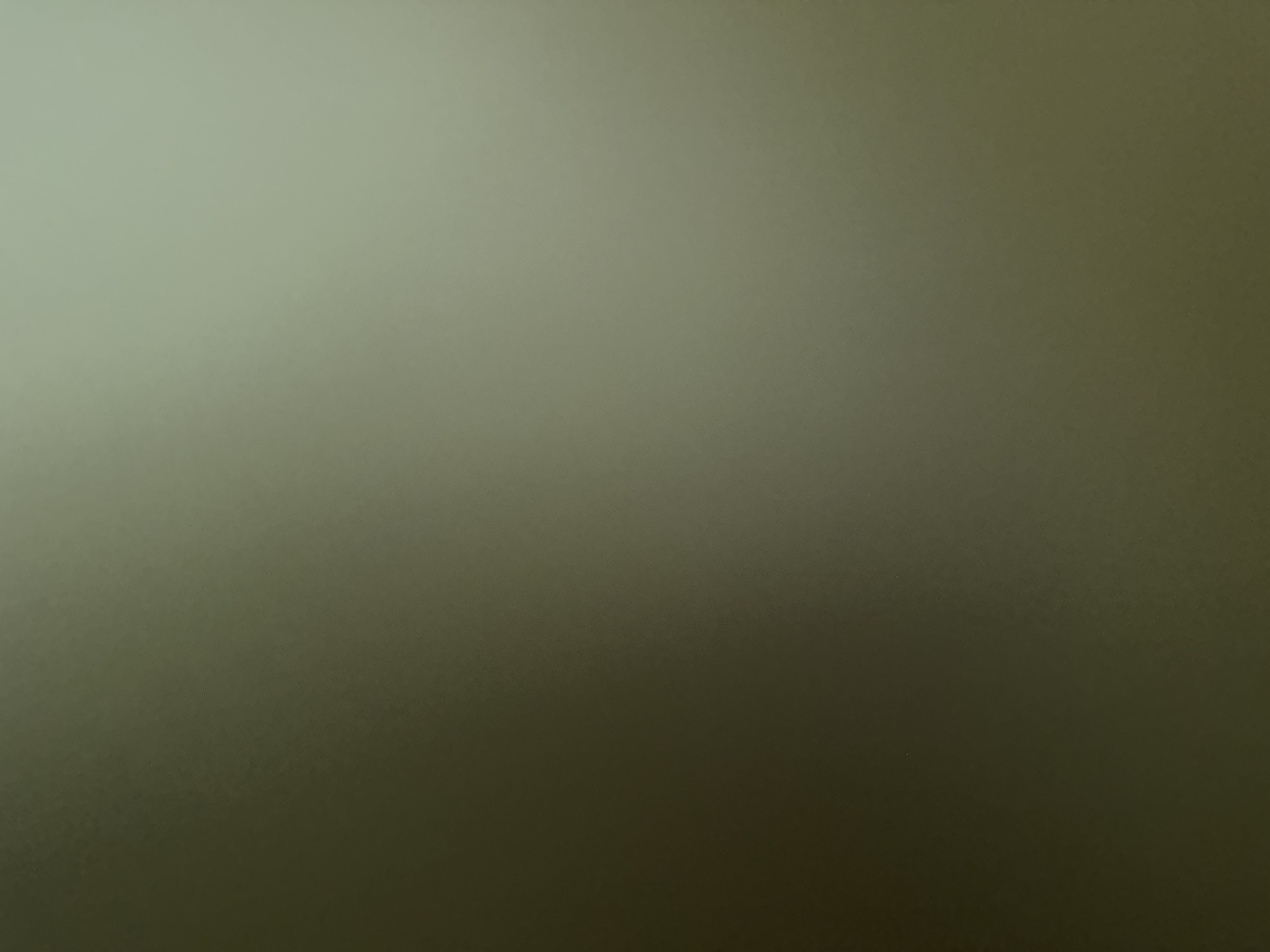 The image is a close-up, abstract shot of a textured surface, likely a wall or fabric, with a gradient of colors ranging from a light, pale green in the upper left to a darker, muted olive green in the lower right. The texture appears granular or slightly speckled, creating a subtle visual noise across the entire frame. There are no discernible subjects, objects, or scenes depicted. No activity or event is taking place, and there are no visible location cues, time of day, weather, or emotional expressions. The image is a study in color and texture, with the light source seemingly coming from the upper left, casting a soft illumination that gradually fades. No text is visible in the image.