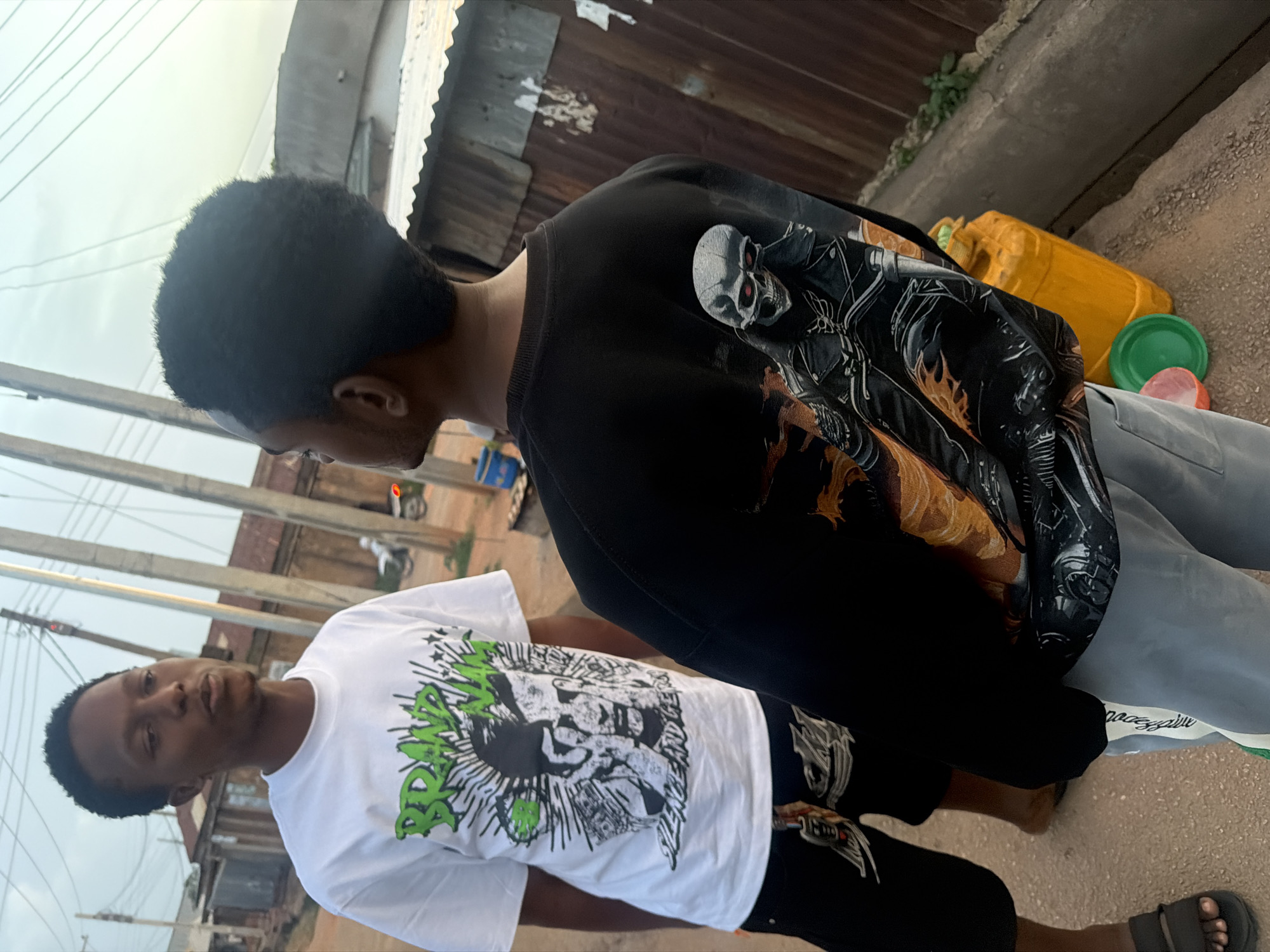 Two young men stand in what appears to be an outdoor urban environment. The man on the left wears a white t-shirt with a graphic print and dark shorts. The man on the right wears a black sweatshirt with a skull graphic on the back and light grey sweatpants. Behind them, a corrugated metal fence is visible, and to the right, a yellow plastic jerrycan sits on the ground. The ground appears to be paved or a dirt path. The overall scene suggests a casual outdoor setting in a potentially developing urban area. The image is captured in Bukuru, Nigeria.