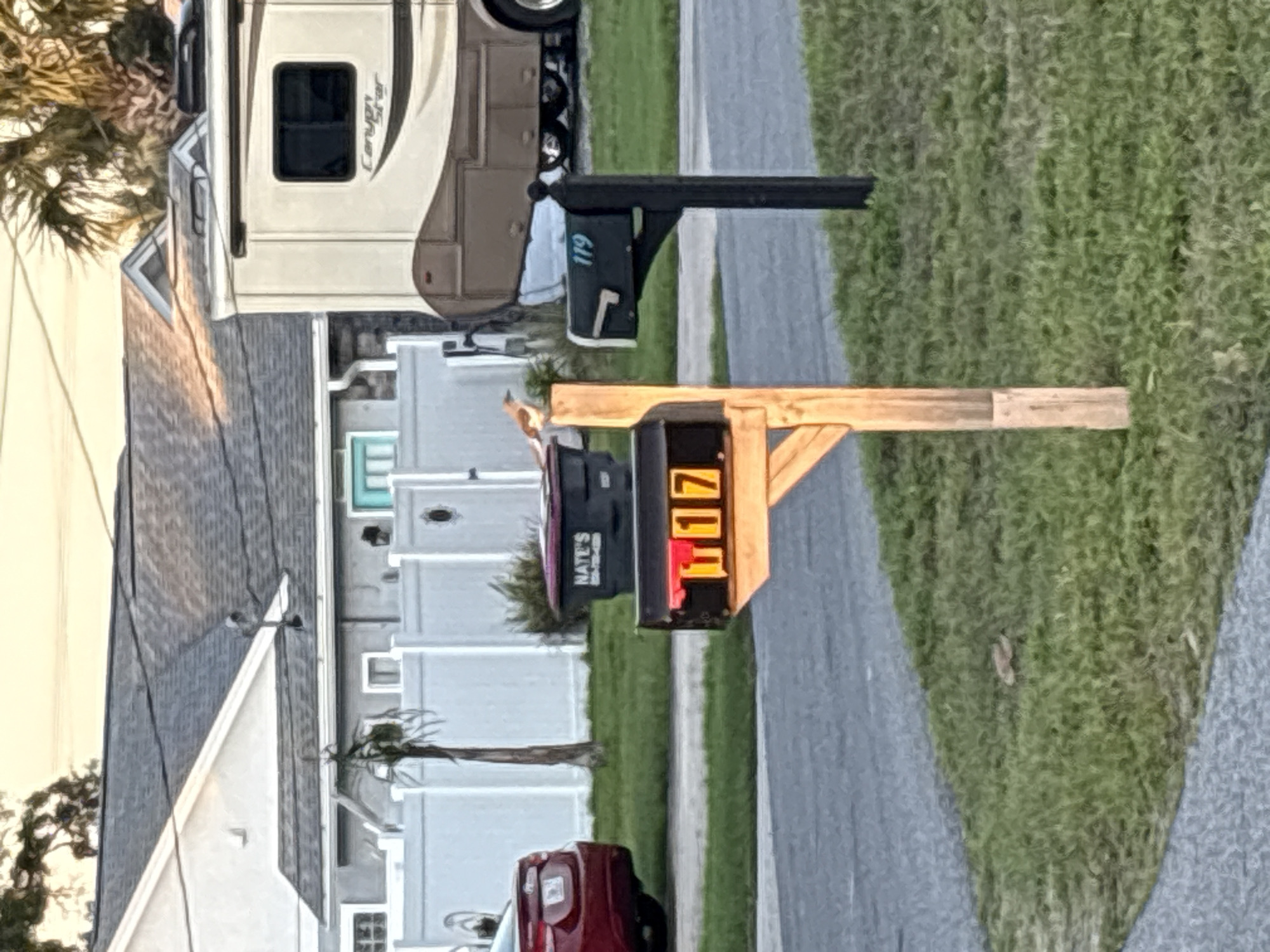 The scene is in Panama City Beach, Florida. An address display box reads "117" in bright orange LEDs. A standard mailbox with the number "119" printed in white is adjacent to the address display. The road in the foreground has a strip of green grass on the right. Behind the mailboxes is a row of white houses with a red car parked in front of one. An RV is also parked along the street.