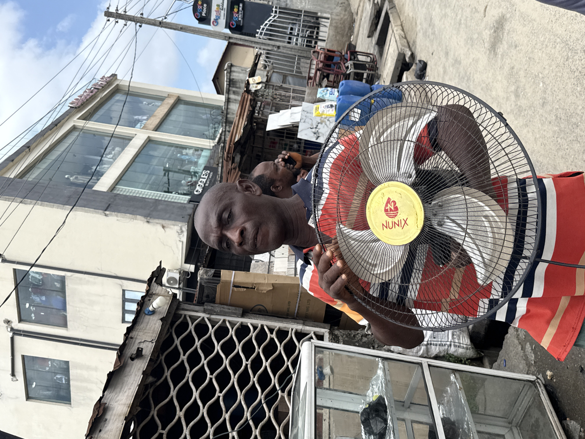 A man holds a "Nunix" brand fan in front of him on a street in Ado, Nigeria. The man is dark-skinned with a shaved head, wearing a striped shirt of red, white, blue, and orange. The fan is black with silver blades and a yellow center. Behind the man, another man is visible, using a phone. The background includes buildings, power lines, a storefront labeled "Giggles Place" in English and another language, and various objects and materials that suggest a commercial or market area.
