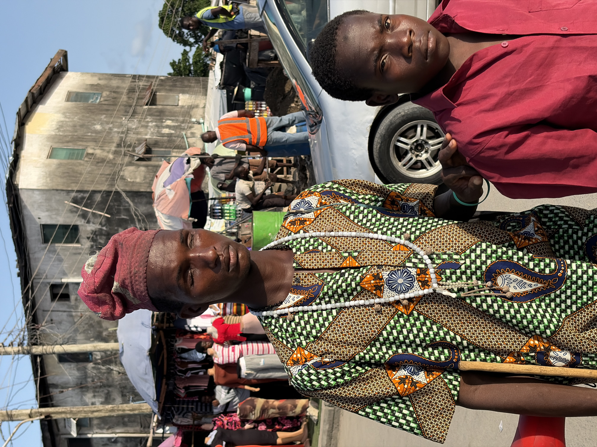 The image is of two dark-skinned individuals in Igboefon, Nigeria. The foreground subject, wearing a red head wrap and patterned green, brown, and orange shirt with a white beaded necklace, holds a wooden cane. Behind and to the right is another person with a maroon shirt. A gray car occupies part of the background, along with several people and a building with weathered walls and visible utility lines. A market stall with clothes is on the left. People are positioned in a crowd around the car.
