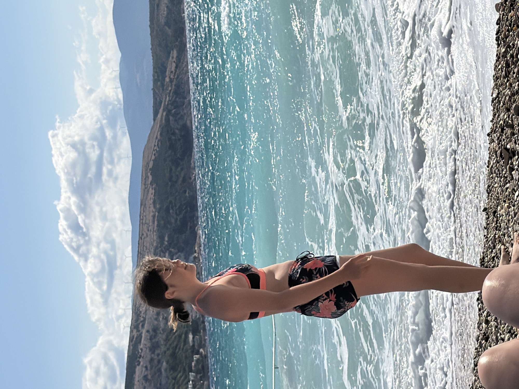 The image depicts a young person standing on a pebbled beach in Općina Baška, Croatia, facing the turquoise sea under a bright, cloudy sky. The person is wearing a floral-patterned bikini and shorts. The sea is choppy with white waves crashing onto the shore. In the background, hills with sparse vegetation and distant wind turbines are visible. The lower portion of the image shows parts of two other people's legs in the foreground, also on the beach. The scene suggests a sunny day at the seaside.