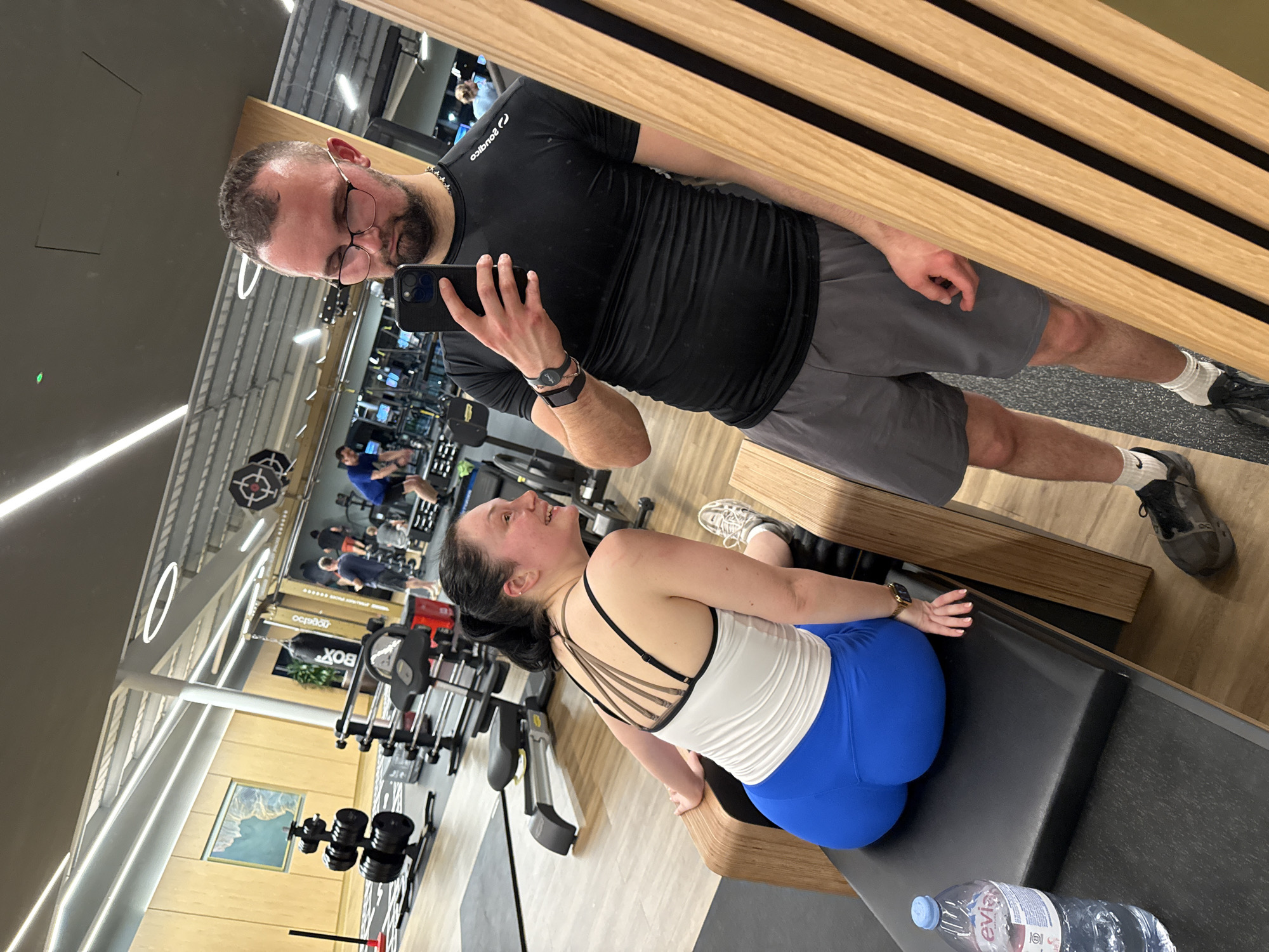 A man and a woman are captured in a mirrored selfie taken in what appears to be a gym setting. The man, on the right of the frame, holds the smartphone and looks down towards the woman with glasses on. He is wearing a black t-shirt, grey shorts, white socks, and dark athletic shoes. The woman, sitting on a black padded bench, looks up towards the man, smiling. She is wearing a white tank top with black straps and blue athletic leggings. A water bottle is on the bench next to her.
In the background of the gym, there are other gym patrons and various workout machines, including weightlifting equipment and rowing machines. The walls are a combination of wood paneling and beige paint. Artificial lighting is provided by overhead lights.
The location is in West Suffolk, United Kingdom.