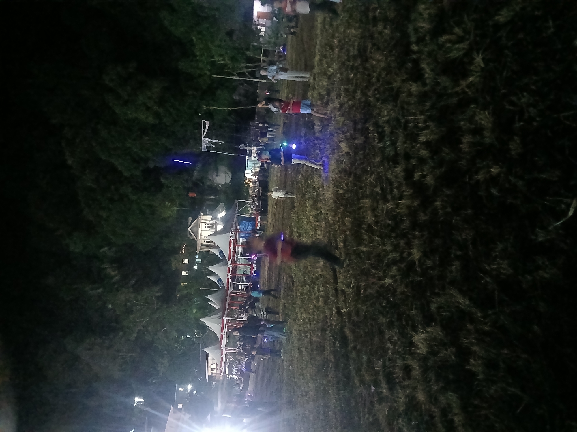 This image depicts a nighttime outdoor gathering in Terempa, Indonesia, possibly a festival or community event. Several tents with white canopies and red trim are set up in the background, suggesting stalls or event spaces. Buildings with illuminated windows are visible behind the tents, nestled among dark trees on a hillside. The foreground is a grassy field, dimly lit by artificial lights, with dry patches of grass and some scattered debris. A group of people, including adults and children, are scattered across the field. Some individuals appear to be walking, while others are standing or interacting. In the mid-ground, several children are holding glowing objects, possibly toys or light sticks, emitting blue light. One child in the foreground is in motion, appearing to run. The overall atmosphere is festive and social, set against a backdrop of natural darkness and illuminated structures.
