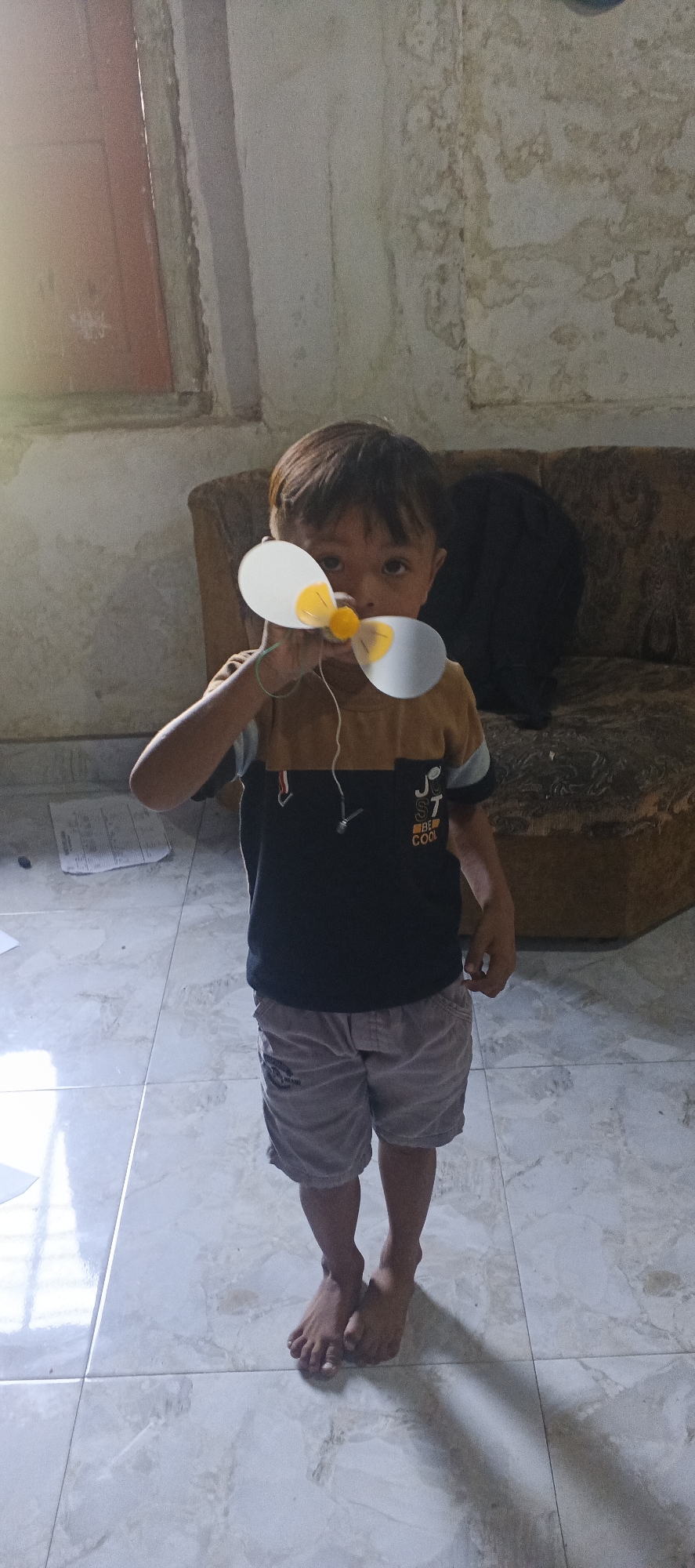 A young child, likely a boy, stands indoors, holding a homemade toy airplane propeller to his face, obscuring his eyes. The propeller has two white blades with yellow centers, connected to a central yellow hub. The child is wearing a brown and black t-shirt with the words "JUST BE COOL" printed on it, and grey shorts. He is barefoot, standing on a tiled floor.

The background shows a worn, textured wall, possibly plastered and painted, with some staining and discoloration. To the left of the child, a section of a red door or window frame is visible. Behind him, a brown, upholstered sofa is partially visible, suggesting a living area. The lighting suggests it might be daytime, with light coming from a window or opening on the left, casting shadows on the floor. The overall scene depicts a candid moment of a child playing with a toy in what appears to be a modest, residential setting.
