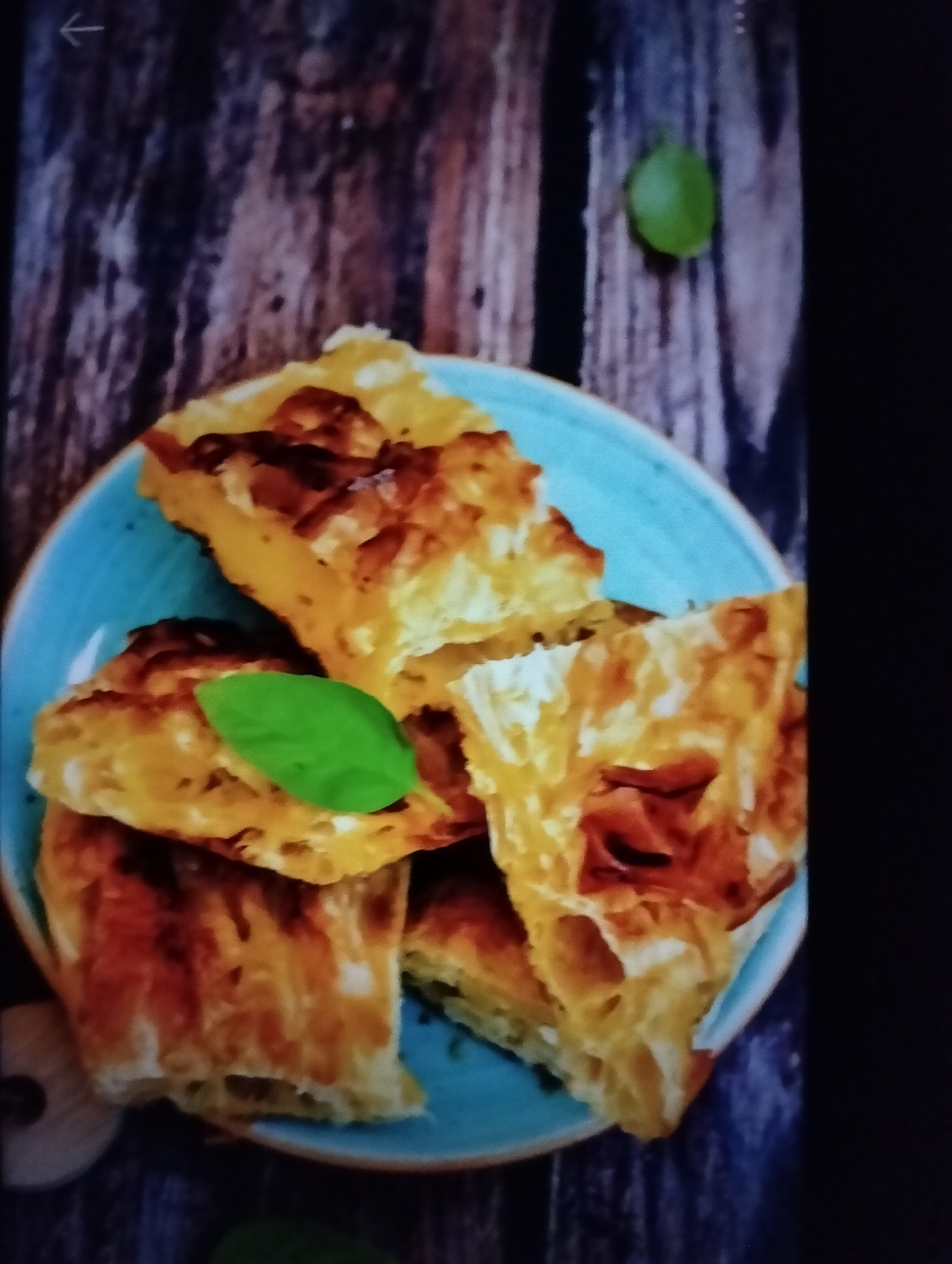 A close-up shot shows slices of golden-brown pastry on a blue plate. The pastry appears layered and possibly filled, with some visible charring on the edges, indicating it has been baked or fried. Two green basil leaves are placed on top of the pastry, one larger leaf as a garnish and a smaller one near the edge of the plate. The plate rests on a dark wooden surface, possibly a table or cutting board, with visible wood grain. In the upper right corner of the wooden surface, a single, small green leaf is also present, possibly a decorative element or part of the same garnish. A faint arrow symbol is visible in the top left corner of the image, pointing to the left. The scene is lit from above, casting shadows that highlight the texture of the pastry and wood. There is no indication of the location being in Payagaboh, Indonesia.