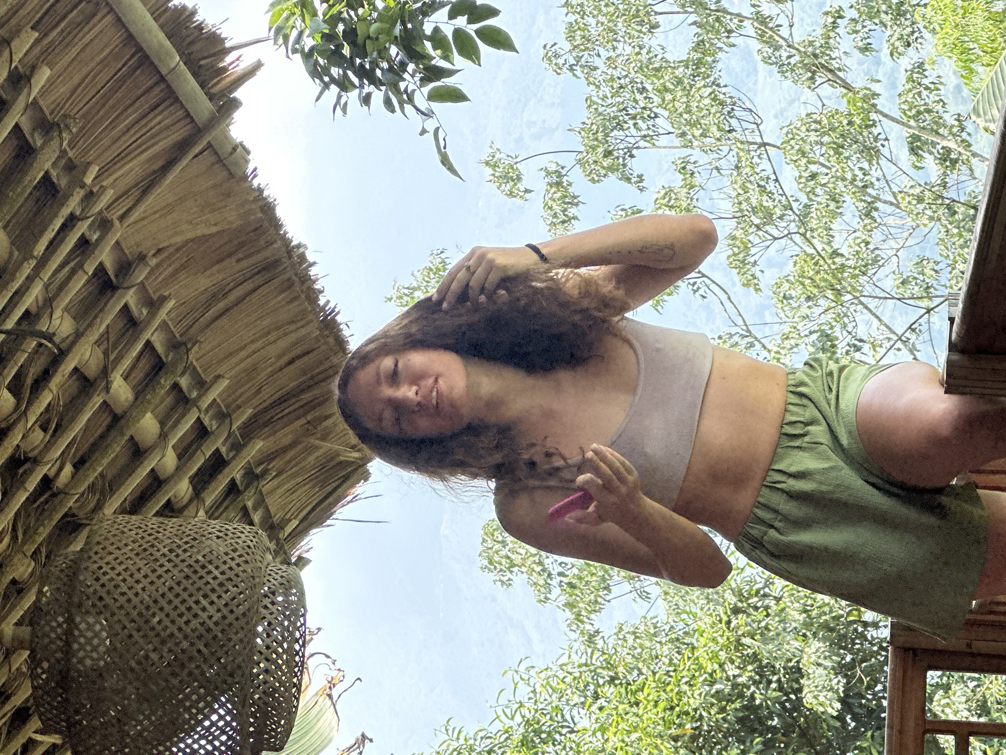 A young woman with long, curly brown hair is the central subject of this low-angle photograph, looking down towards the camera with a gentle smile. She wears a light-colored crop top or sports bra and olive green shorts with an elasticated waist. Her left hand is near her hair, while her right hand holds a small, pink, cylindrical object. A dark bracelet adorns her right wrist, and a ring is visible on her left hand.
To the left of the frame, a rustic thatched roof structure, constructed from bamboo poles and dried plant materials, extends diagonally. Hanging from this structure is a round, woven basket-like object with an open lattice pattern, possibly a lamp shade or decorative element.
The background is dominated by a bright, clear blue sky with some faint, wispy clouds. Lush green foliage from numerous trees fills the upper right and lower right portions of the image, suggesting a vibrant, natural outdoor environment. The bright lighting indicates it is daytime, under sunny weather conditions. The overall setting is natural and rural, consistent with a location like Thông Nguyên Commune, Vietnam. No visible text is present in the image. The woman appears relaxed and content.