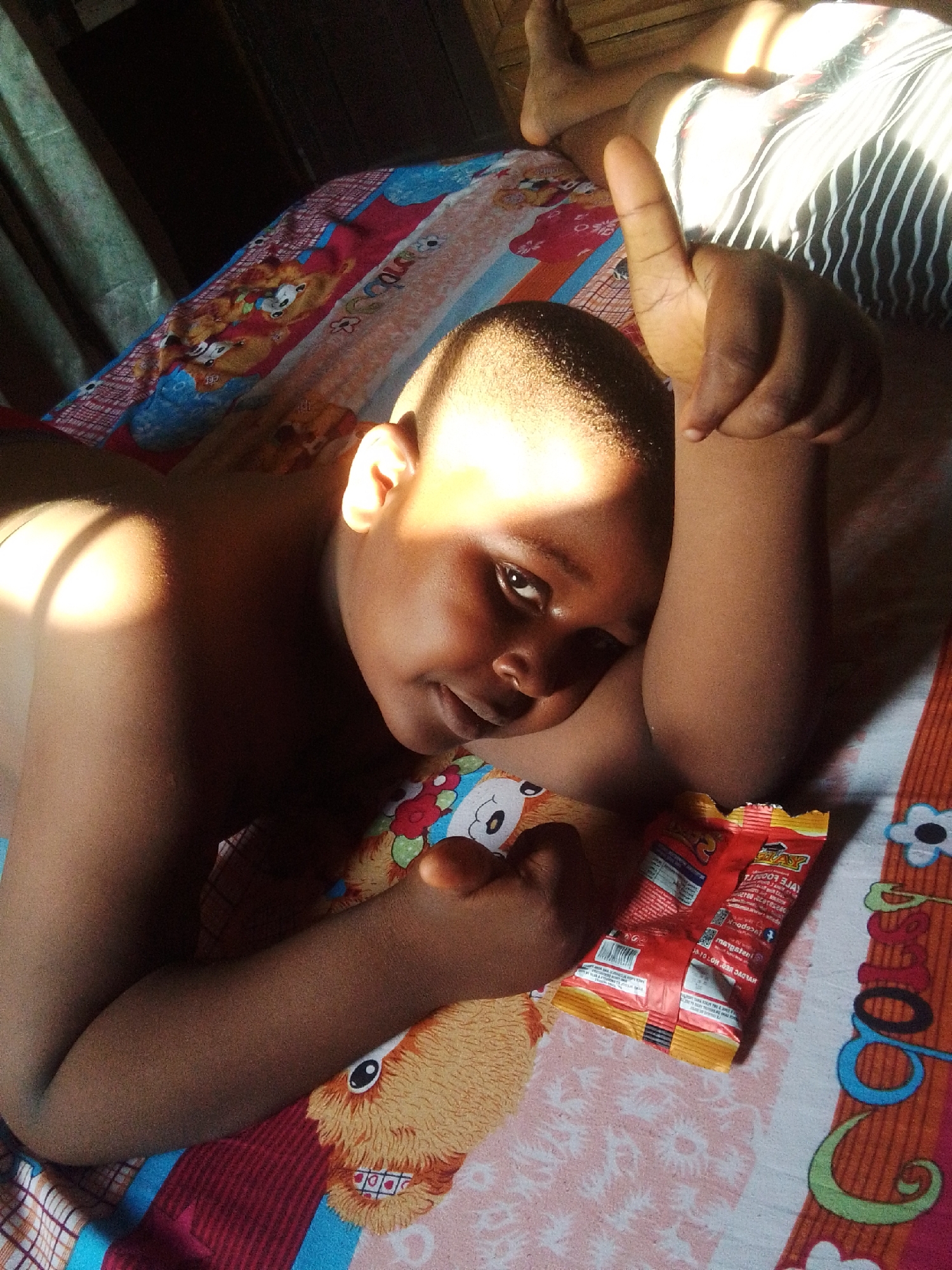 The image shows a child lying on a bed with a colorful blanket. The child is looking towards the camera with a smile and has their right arm extended upwards, with their thumb pointing up. A snack packet is lying on the bed next to the child's arm. The room appears to be illuminated by natural light, casting shadows on the wall and bed. The blanket has cartoon characters printed on it. The child is wearing a black and white striped garment. The environment suggests a home setting, likely in a bedroom. The child's pose and the snack packet imply a casual and relaxed moment. The image does not provide specific details that can definitively place the location in Nkpor, Nigeria.