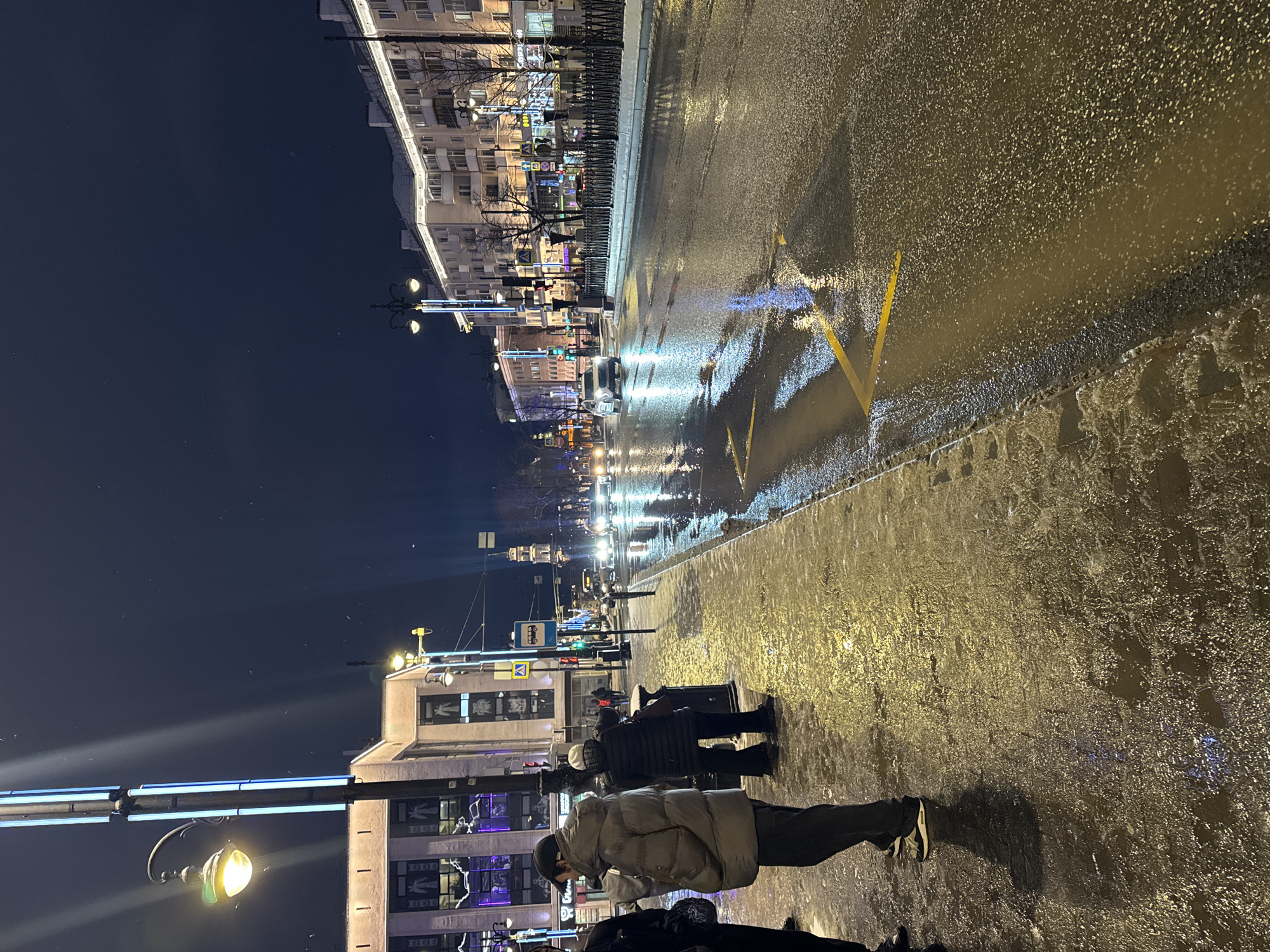 A vertical, night-time photograph captures a wet city street in Perm, Russia. The scene depicts a bustling urban environment illuminated by artificial light sources.
In the foreground and mid-ground on the right, a wide sidewalk, glistening with moisture, hosts several pedestrians. Two individuals in heavy winter coats and hats stand prominently near a dark trash bin. Another person is partially visible in the bottom right corner. The sidewalk transitions into a wet asphalt road on the left, reflecting bright streaky lights from cars and streetlamps. A distinct yellow arrow road marking is visible on the asphalt, pointing towards the top left.
Further down the street, cars with illuminated headlights and taillights create bright reflections on the dark, wet road surface, indicating ongoing traffic. A metal fence lines the edge of the road, separating it from the buildings beyond. On the far left, a tall streetlight with a bright yellow glow casts light onto the lower left corner of the frame.
Along the left side of the street, multi-story buildings with numerous windows are brightly lit, their facades showcasing various commercial signs and decorative lighting. Above the buildings, the night sky is deep navy blue. A blue bus stop sign with a white bus icon and a small yellow triangular sign with an exclamation mark are visible in the mid-ground, indicating a public transport stop. The overall impression is of a cold, damp, but vibrantly lit winter night in a city. The wet surfaces and numerous reflections are a prominent visual feature of the image. No discernible text is clearly readable on the buildings or signs beyond the basic icons.
