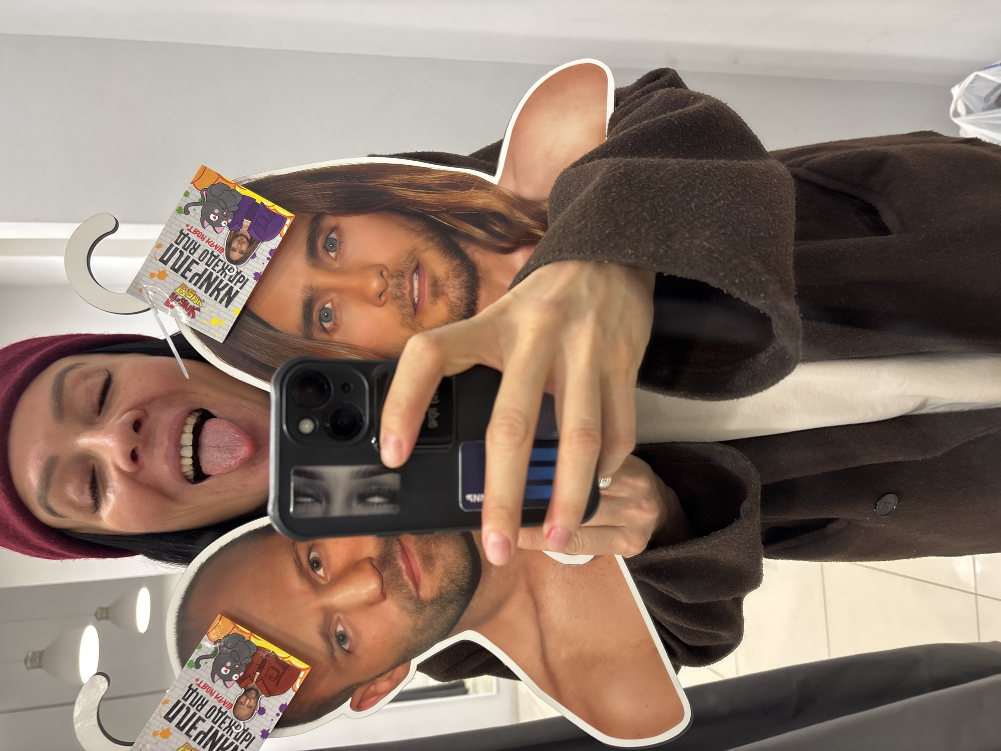 A woman with a maroon hat and open mouth is taking a selfie with a person holding a phone in front of a mirror, which is reflecting the phone, two cutout heads of male celebrities and a brown coat hanging on a hook. The cutout heads have tags that read "LEON KLIMOV OLYANKA". The location appears to be in a clothing store, possibly in Chaikovsky, Russia.