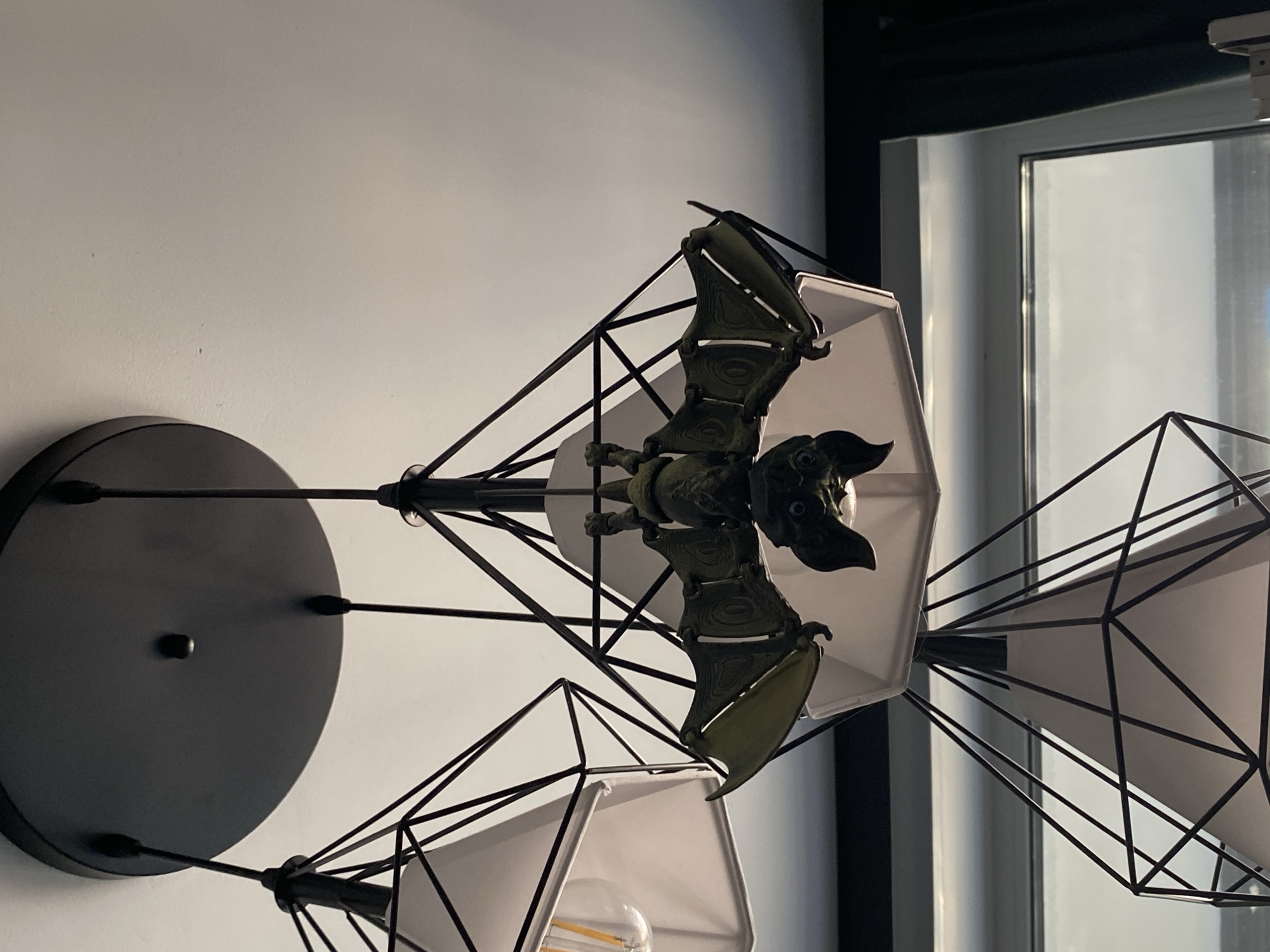 This is a close-up, low-angle shot of a ceiling light fixture. The fixture has a black circular canopy attached to the ceiling, from which three black metal geometric pendants hang. Each pendant contains a white conical lampshade. One of the pendants is adorned with a decorative, dark green, bat-shaped ornament, hanging upside down. The bat has outstretched wings and appears to be made of plastic or resin. The lighting suggests an indoor environment, possibly a room with a window visible in the background, which shows a cloudy sky. The image does not provide any specific visual cues to indicate its location in Yekaterinburg, Russia.