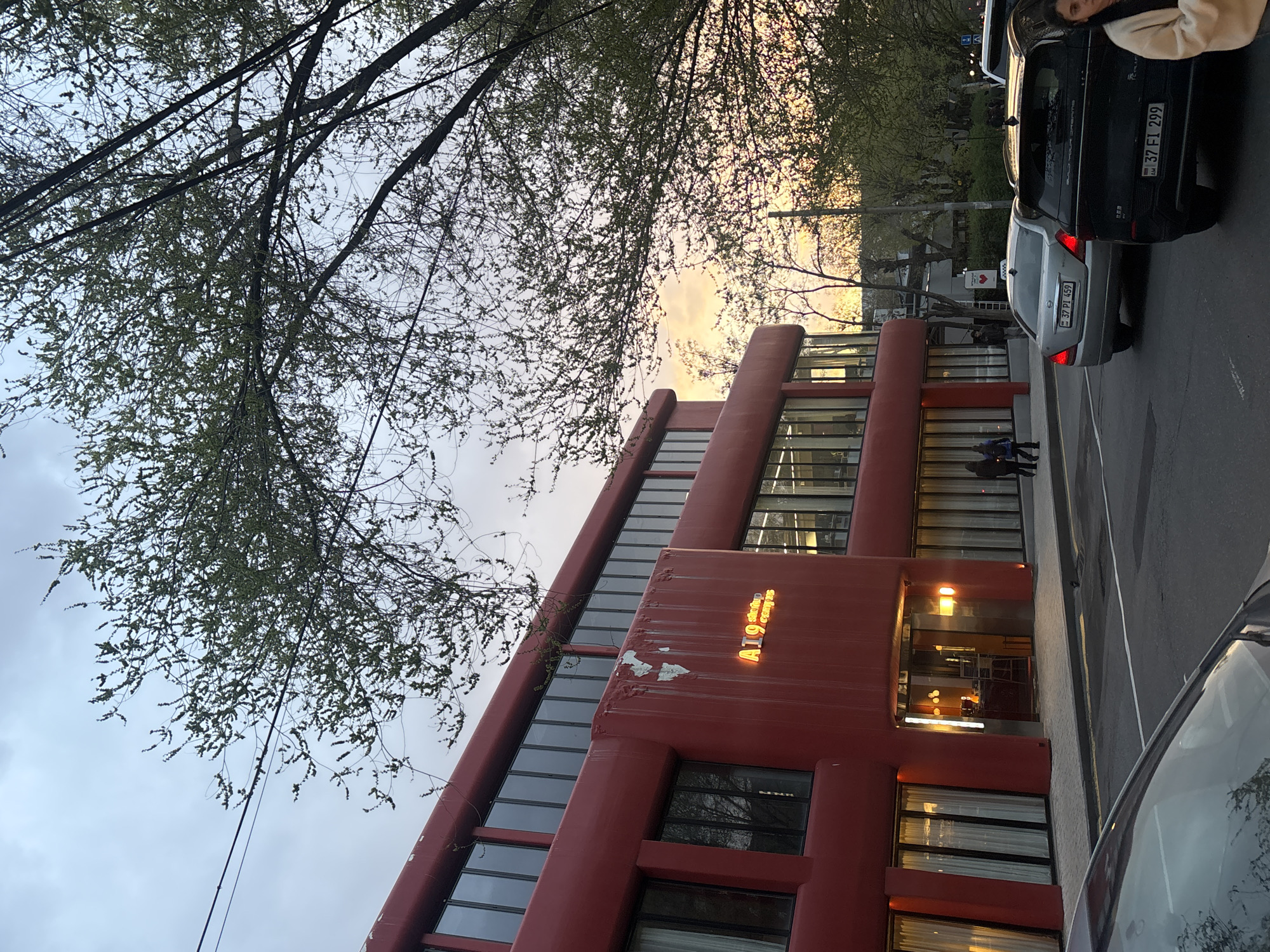 The image shows a multi-story, red building with glass windows. A sign on the building reads "A19 Student Campus". The sky is visible above the building, with a mix of clouds and a hint of sunset colors. There are trees with sparse foliage in the foreground. Several parked cars are visible on the street adjacent to the building. A person is partially visible next to the car on the upper right corner of the image. A few people stand on the sidewalk in front of the building, but they are far away and not clearly defined. The license plate on the car closest to the building reads "37 PI 459" and the other reads "37 EI 299". The location is Yerevan, Armenia.