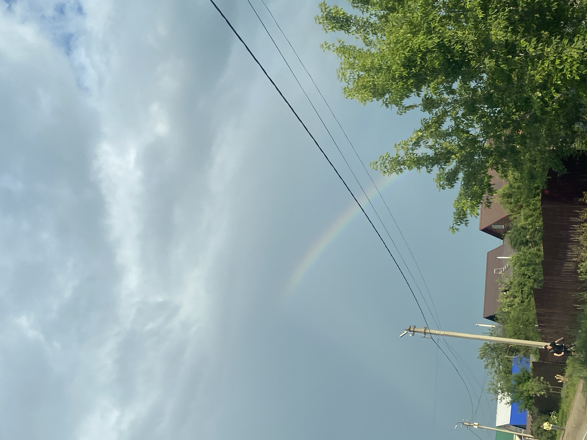 The image depicts a cloudy sky with a faint rainbow visible in the upper left quadrant. In the foreground, there is a row of houses with brown roofs and a wooden fence. A large, leafy green tree is on the right side of the image. Two power poles are visible, one in the center and another to the left. On the left side of the image, a person in a yellow shirt is walking, and a person on a bicycle is in the center foreground. The scene appears to be a residential area.