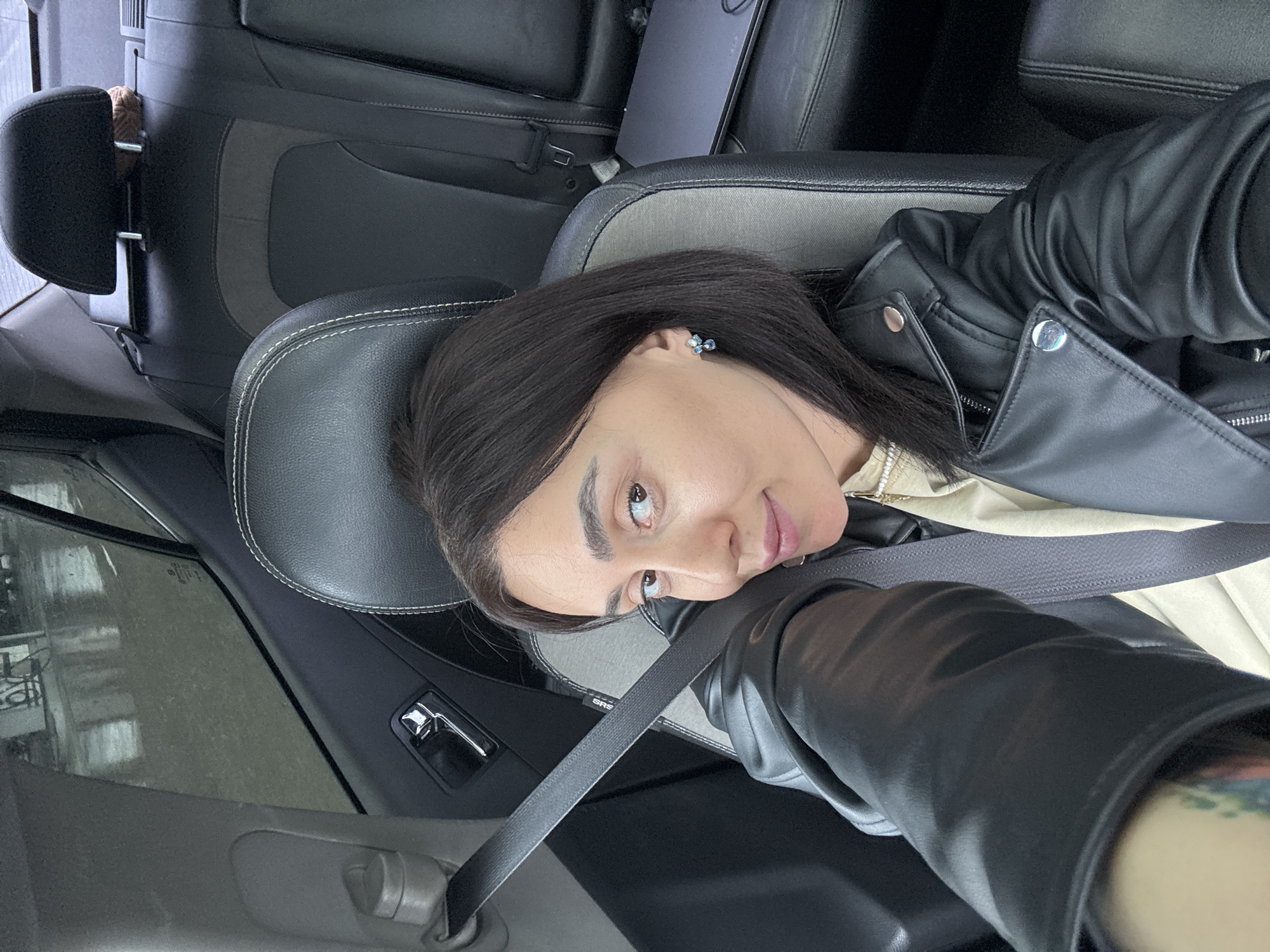 A female individual is seated in the front passenger seat of a vehicle, taking a selfie. She has dark hair, a light complexion, and is wearing a black faux leather jacket over a light-colored, possibly beige or cream, top. A black seatbelt is fastened across her chest. Small, blue-toned stud earrings are visible on her right ear. A multi-colored tattoo is partially visible on her left forearm. The car interior features black and grey two-tone upholstery, with a dark-colored laptop resting on the rear passenger seat. Chrome door hardware is visible on the driver's side door panel. The exterior environment, visible through the driver's side rear window, shows a wet, reflective ground surface under an overcast sky, suggesting recent precipitation. The scene is located in Tbilisi, Georgia.
