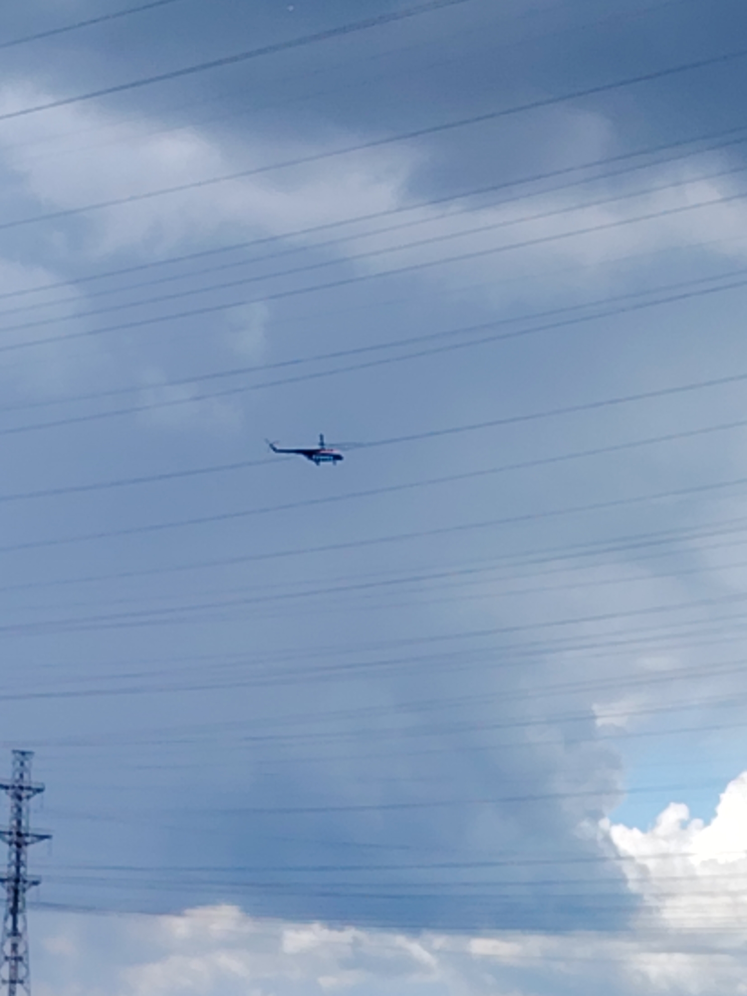 The image depicts a helicopter in flight against a cloudy sky. Power lines are visible in the foreground and run across the image, and an electricity pylon can be seen on the lower left. The location appears to be Chaikovsky, Russia, given the presence of power lines and a helicopter in an outdoor setting.