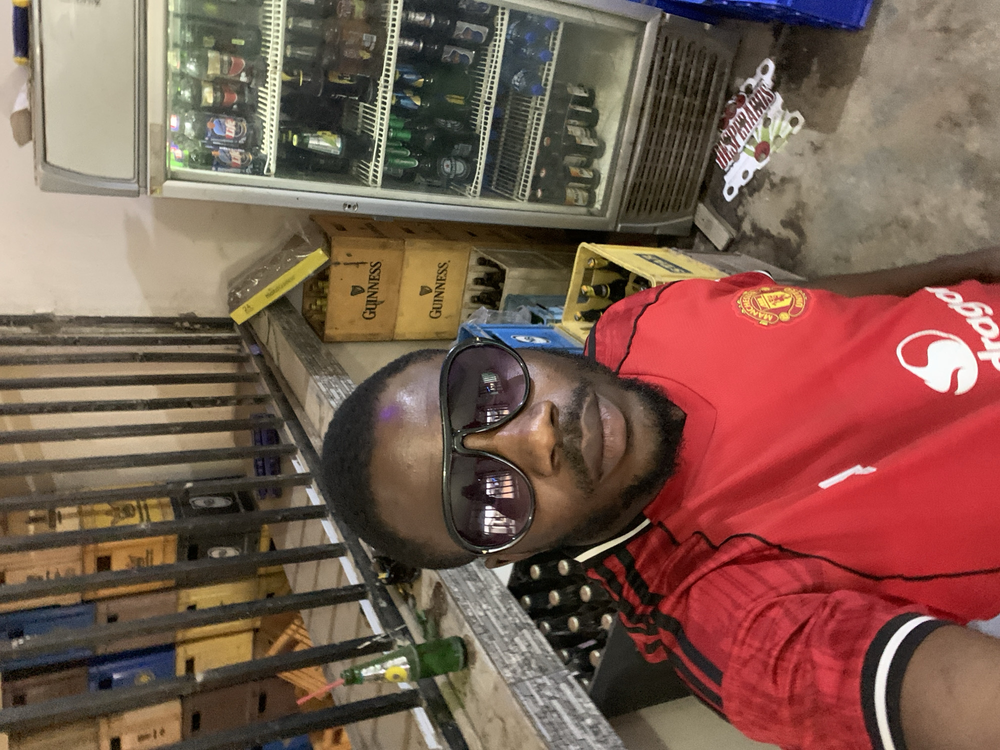 A selfie shows a man in sunglasses and a red Manchester United shirt in New Stadium, Nigeria. Behind him is a refrigerated display case filled with beverages, several stacked crates of beer (some Guinness branded), and scattered Desperados promotional items. He is positioned near a railing.