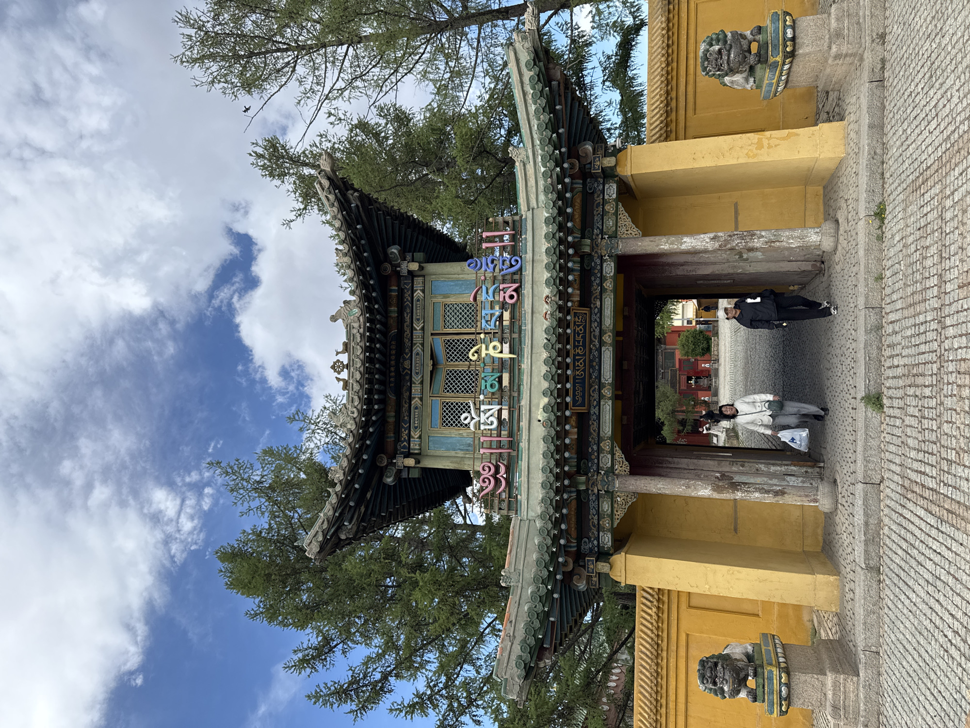 The image depicts the entrance to a temple in Ulaanbaatar, Mongolia. The ornate gate features traditional Mongolian architecture, including a tiered roof, decorative carvings, and colorful inscriptions in Mongolian script. The structure is painted in yellow, with dark brown and teal accents. Two people are walking through the entrance. The ground is paved with grey bricks. The sky is blue with scattered clouds, and trees flank the gate. Two guardian lion statues sit atop pedestals, flanking the pathway.