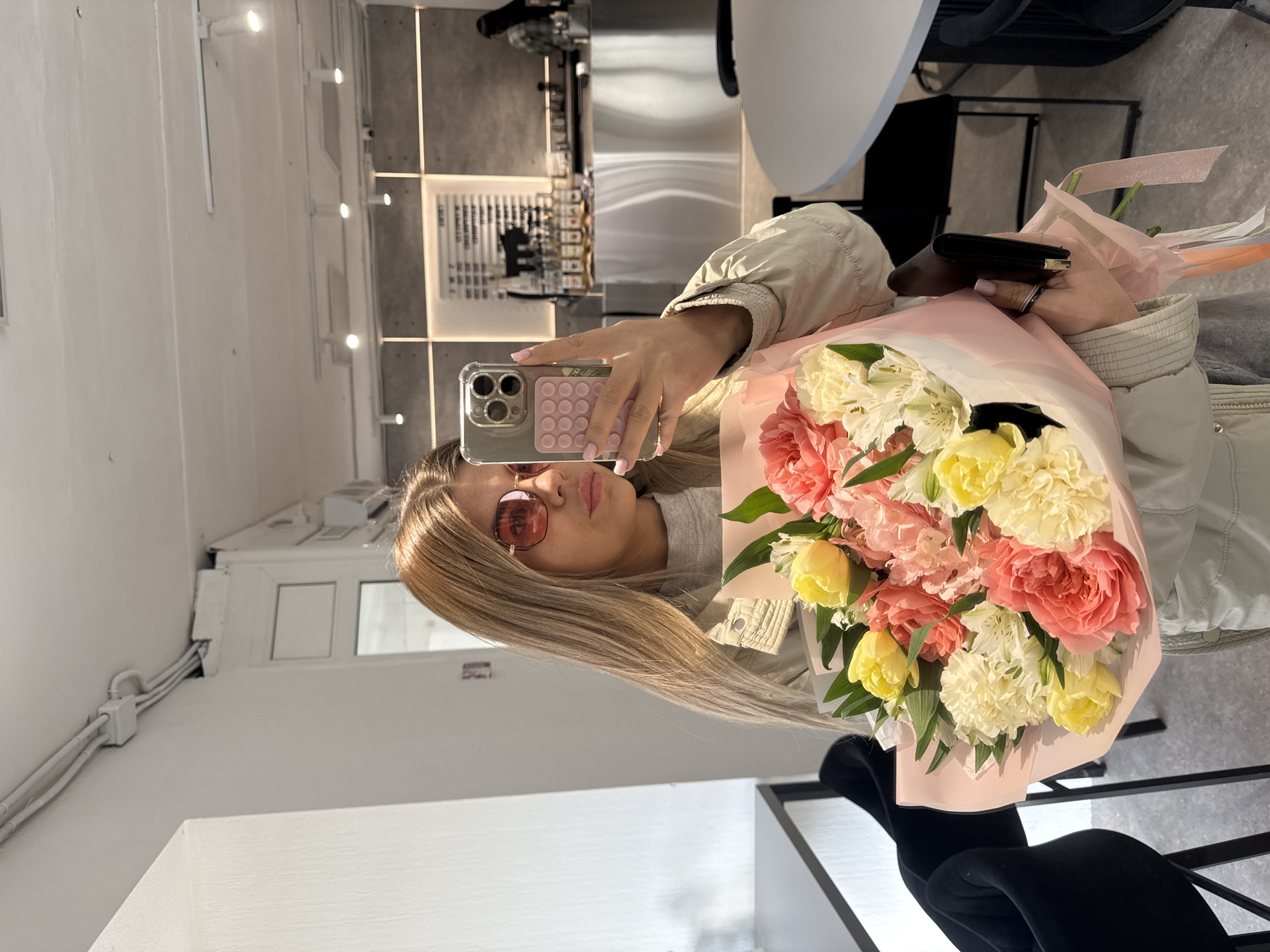 A young woman is taking a selfie indoors, likely in a cafe or shop in Chaikovsky, Russia, given the context. She is holding a large bouquet of flowers, primarily consisting of yellow tulips and pink and white carnations, wrapped in pink paper. She is wearing reddish-brown sunglasses and a light-colored jacket. The phone she is using to take the picture has a pop socket on the back. The background reveals a modern interior with neutral tones, featuring a tiled wall, light fixtures, and what appears to be a display case. The environment suggests a stylish and possibly upscale establishment.