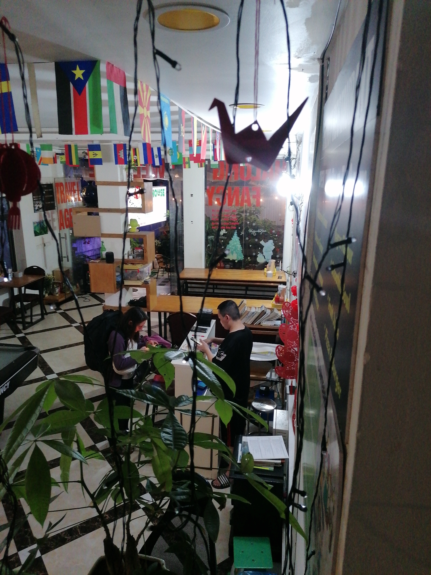 This image captures an indoor scene, likely a common area of a hostel, cafe, or travel agency, given its decor and activity. The room features a checkered tile floor and a white ceiling, with some visible discoloration. A defining characteristic is the string of numerous small flags from various countries, including South Sudan, United Arab Emirates, and North Macedonia, hung across the ceiling, suggesting a theme catering to international travelers. Red lantern-like decorations and a dark origami crane also hang from above, contributing to the eclectic atmosphere.
In the foreground, a large, leafy green plant partially obstructs the view, and string lights are draped, adding a festive glow. Two individuals are present: on the left, a person wearing a purple top and a black backpack is seated at a counter, focused on something in front of them, possibly a laptop or paperwork. On the right, another person in a black t-shirt stands behind a counter, also looking down at a laptop or administrative tasks. This activity suggests guest services, travel planning, or casual work.
A large window at the back reveals bright lights from outside, and "MERRY CHRISTMAS" is clearly visible on the glass, accompanied by Christmas tree and snowman decorations. This indicates the photograph was taken during the Christmas season, likely in the evening or at night. Other visible text includes "TRAVEL AGE" (possibly "TRAVEL AGENCY") and "HOUSE" on signs, and "FANCY" on the window. The space is furnished with wooden tables and chairs, and a multi-tiered wooden shelving unit holds various items. The scene portrays a functional and welcoming environment for travelers in Bai Chay Ward, Vietnam.