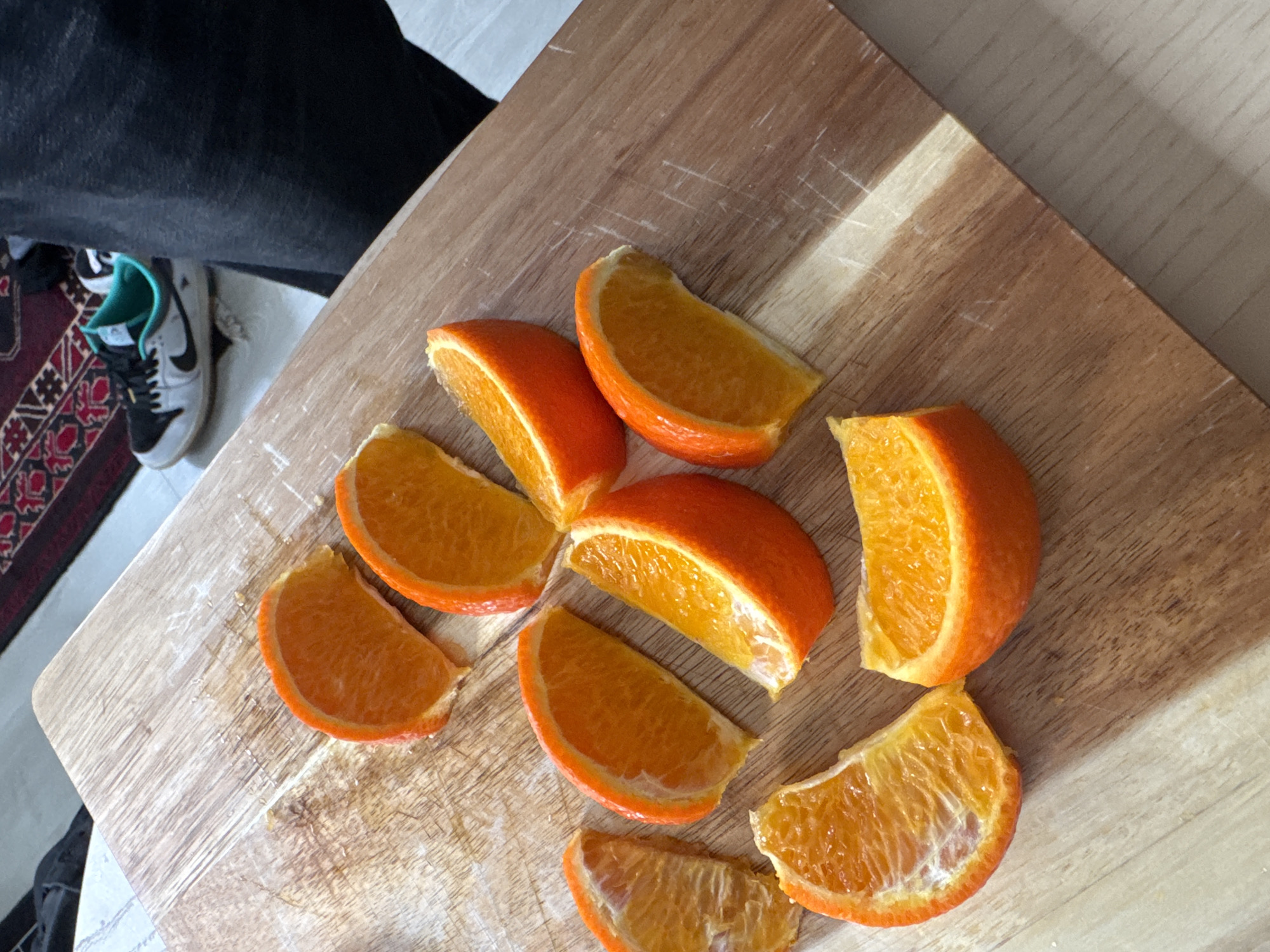 The image shows a wooden cutting board with several orange segments arranged on it. The oranges appear to be freshly cut. To the left of the cutting board, part of a person is visible, wearing dark-colored pants and a gray Nike sneaker with turquoise accents. The scene appears to be indoors, with the edge of a patterned rug visible near the sneaker. The image does not provide any information indicating the scene's location is in New York City or the United States.