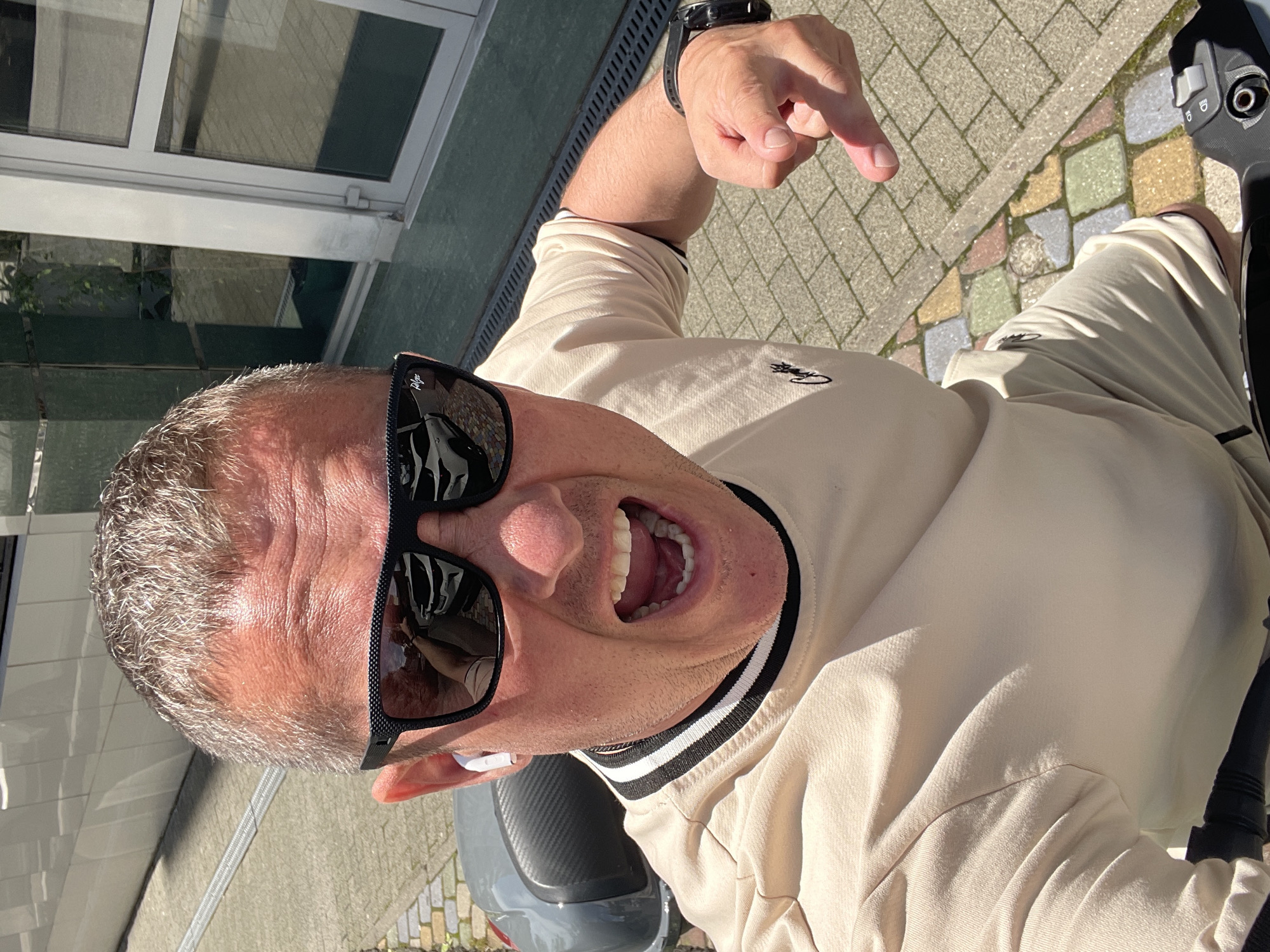 A man with short, graying hair, wearing black sunglasses and a tan Lacoste jacket, is taking a selfie from a high angle. He's pointing with his right hand and has his mouth open. He is positioned on a gray scooter, riding on a paved surface with multi-colored stones. A building is visible in the background, with window reflections. It is in Sochi, Russia.