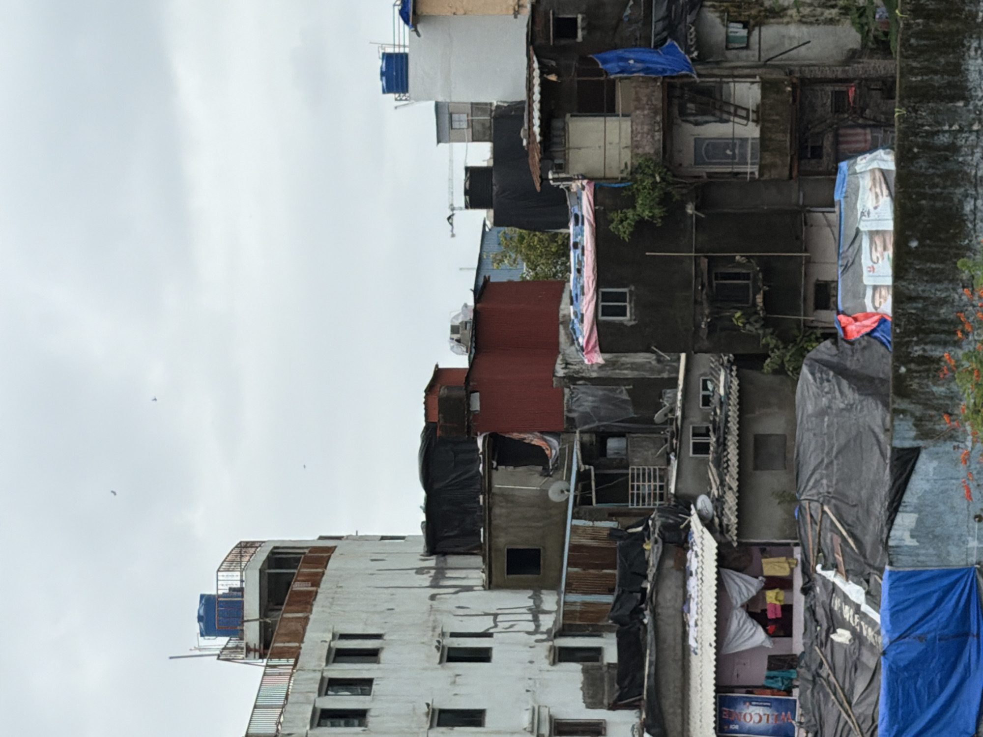 The image depicts a dense urban landscape with a collection of buildings in Mumbai, India. The structures vary in height and construction, showcasing a mix of multi-story concrete buildings and makeshift dwellings. Many buildings are weathered and show signs of age and repair, with some covered by tarps and corrugated metal sheeting. Water tanks are visible on the rooftops of some structures, indicating a common urban utility solution. Laundry hangs from some of the buildings, suggesting residential use. The scene is characterized by its density and the overlapping nature of the constructions, typical of informal settlements or densely populated areas within a large city. The sky is overcast, contributing to a muted color palette. The overall impression is one of a complex, lived-in urban environment.