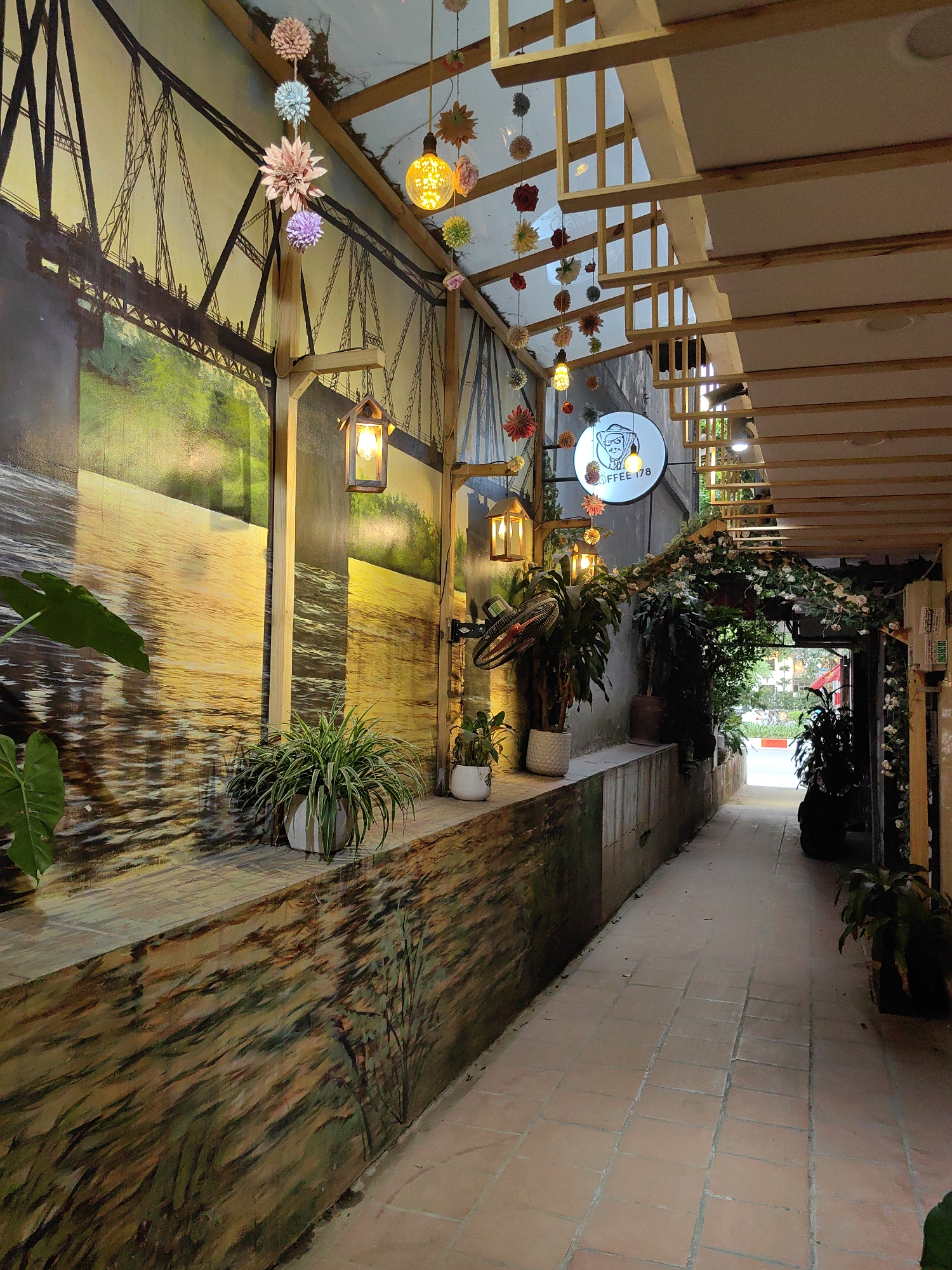 This image captures a narrow, well-decorated outdoor or semi-enclosed walkway, likely leading to a cafe or shop, set in Hà Nội, Vietnam. The pathway features terracotta-colored tiled flooring and is flanked on the left by a low wall and a larger vertical wall, both adorned with murals.
The mural on the left depicts a serene landscape with a large bridge spanning over water, reflecting warm sunlight. The upper part of the mural shows the intricate structure of the bridge with small figures walking across it, while the lower part portrays the shimmering water. Below the bridge mural, the low wall features a painting resembling textured earth or rock. Several potted plants, including a large leafy one in the foreground and a spiky green plant in a white pot, are placed along the low wall, contributing to a lush feel.
The walkway is illuminated by several decorative lights, including hanging spherical bulbs and lantern-style fixtures with warm glows, suggesting it is either evening or an interior space relying on artificial light. Overhead, various colorful pom-pom-like flower decorations are strung across the ceiling and hang down, adding a festive and whimsical touch.
Further down the pathway on the right, a circular white sign with a cartoon-like image of a person wearing a cap and glasses is visible. The text on the sign clearly reads "COFFEE 178". Below the sign, an oscillating fan and more potted plants are present. The archway beyond is heavily draped with artificial vines and white flowers, creating a natural, inviting tunnel leading to what appears to be an open area or street beyond, with a glimpse of red and white road markings. The ceiling structure consists of wooden beams and panels, with some areas appearing to have a clear or translucent covering, allowing some light through. The overall atmosphere is cozy, inviting, and creatively decorated.
