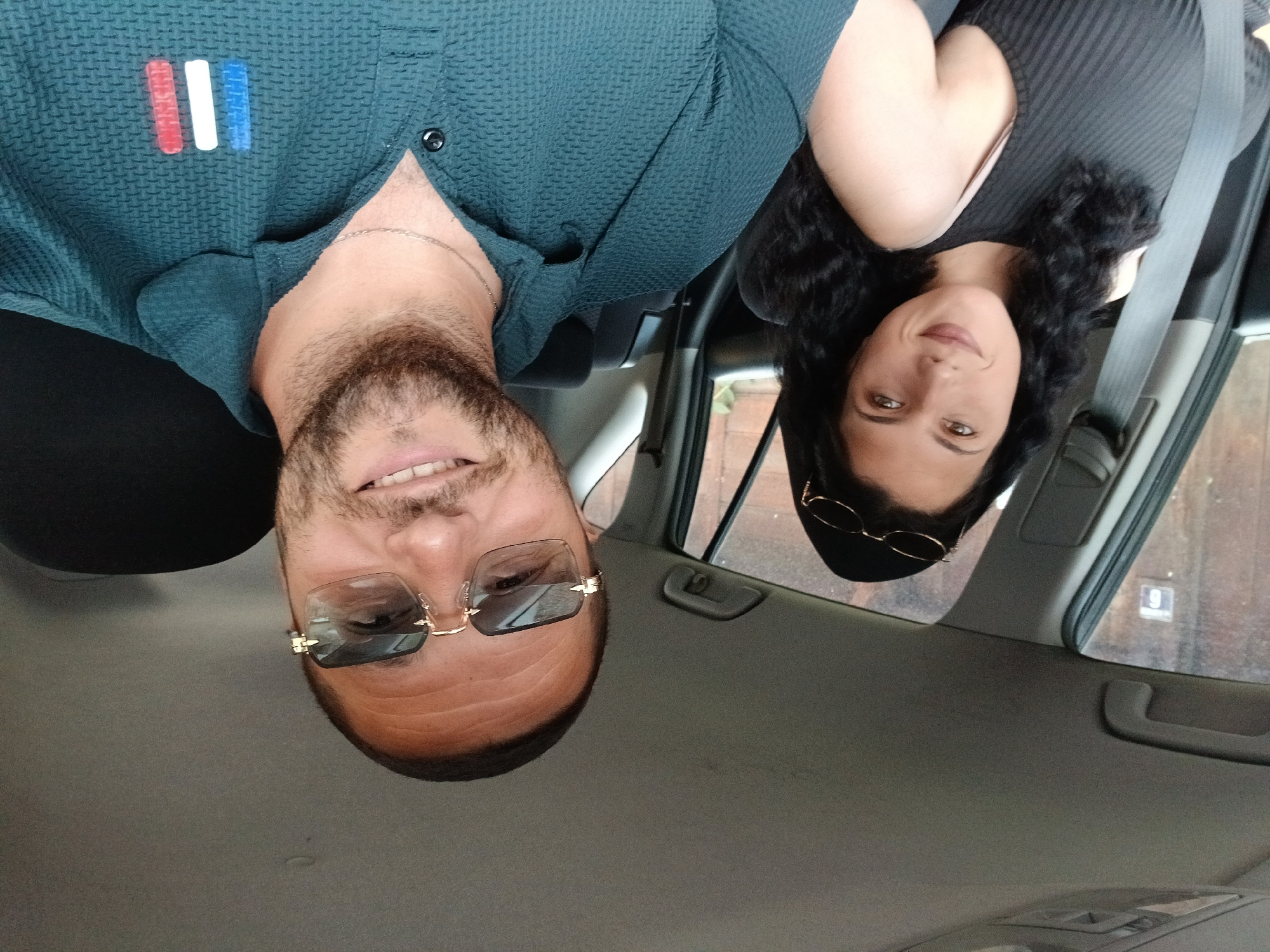 Two individuals are captured in a selfie within the interior of a car. The man, positioned on the left side of the image, sports glasses and has a beard. He's wearing a teal-colored button-down shirt adorned with a small rectangular patch exhibiting the colors of the French flag. To the right, a woman is visible wearing a black top and a seatbelt. The interior of the car, including the dashboard, and window frames are visible. Through the car window, a building with the number "9" on it is visible. This image was taken in Teteven, Bulgaria.