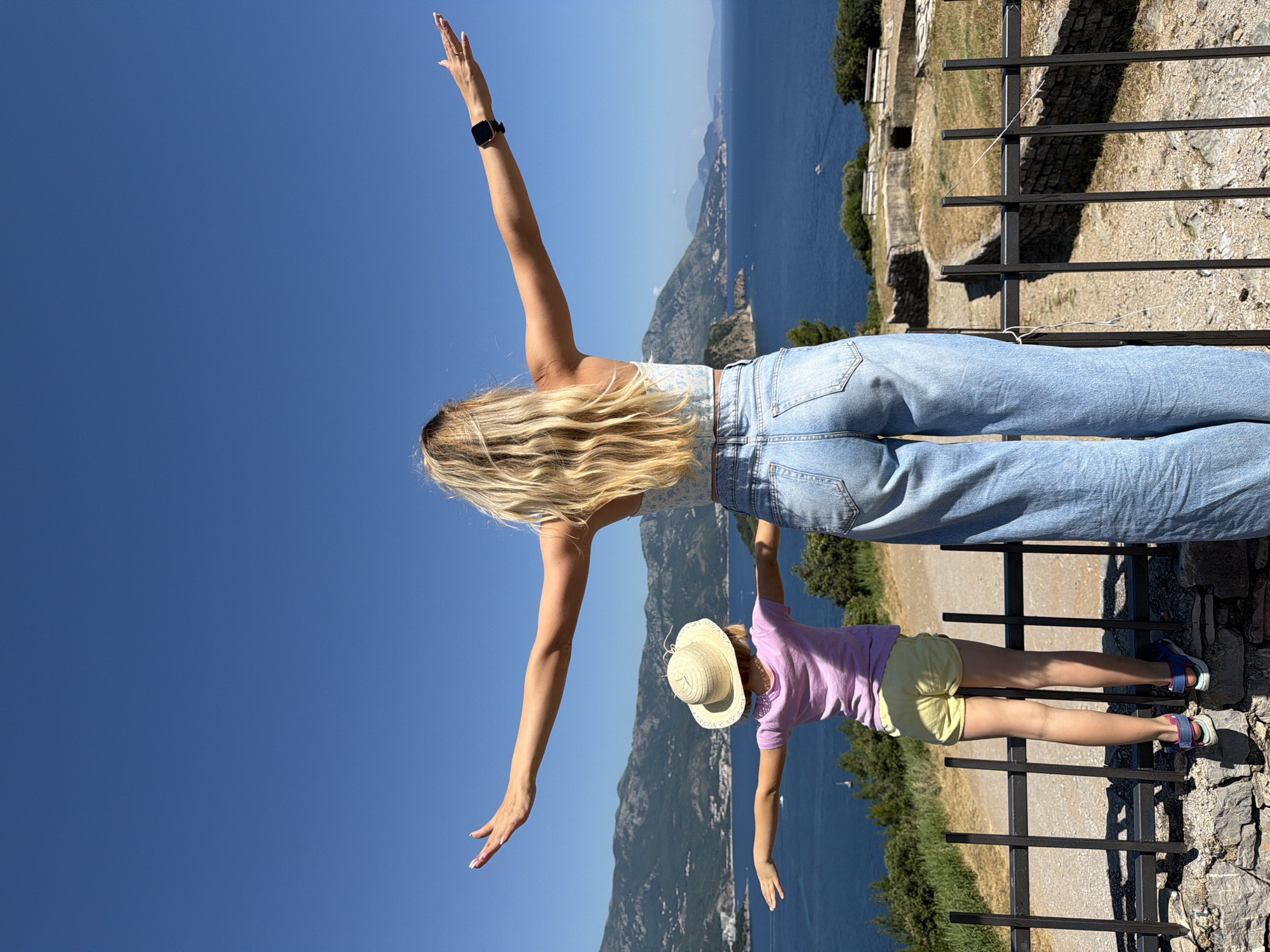 This image presents an elevated panoramic view of the Adriatic Sea coastline in Budva, Montenegro. An adult female and a young child are positioned with their backs to the camera, arms horizontally outstretched, facing the expansive sea and distant mountains under a clear blue sky.
The adult female has long, wavy blonde hair and wears a light-colored, patterned crop top with light blue, wide-leg denim jeans. A black smartwatch is visible on her left wrist, along with a ring on her left ring finger. The child, a young girl, wears a light pink t-shirt, light yellow shorts, and blue and pink athletic sandals. She is also wearing a straw hat. The child stands on a low stone wall directly behind a black metal fence, which serves as a barrier in the immediate foreground.
The fence consists of vertical black metal bars supported by horizontal crossbars. A thin white string or wire is tied across the fence on the right side. Below the fence and the vantage point, a sloped terrain shows patches of dry grass and exposed stone, indicative of an ancient or historical site, possibly a fortification. Two white wooden benches are visible further down the slope.
The deep blue Adriatic Sea occupies the middle ground and horizon, dotted with several small watercraft, including a sailboat and a motorboat. The background is dominated by a range of green, tree-covered mountains that meet the sea, characteristic of the Montenegrin coast. The scene is illuminated by strong sunlight, creating clear shadows and indicating a bright, clear day.