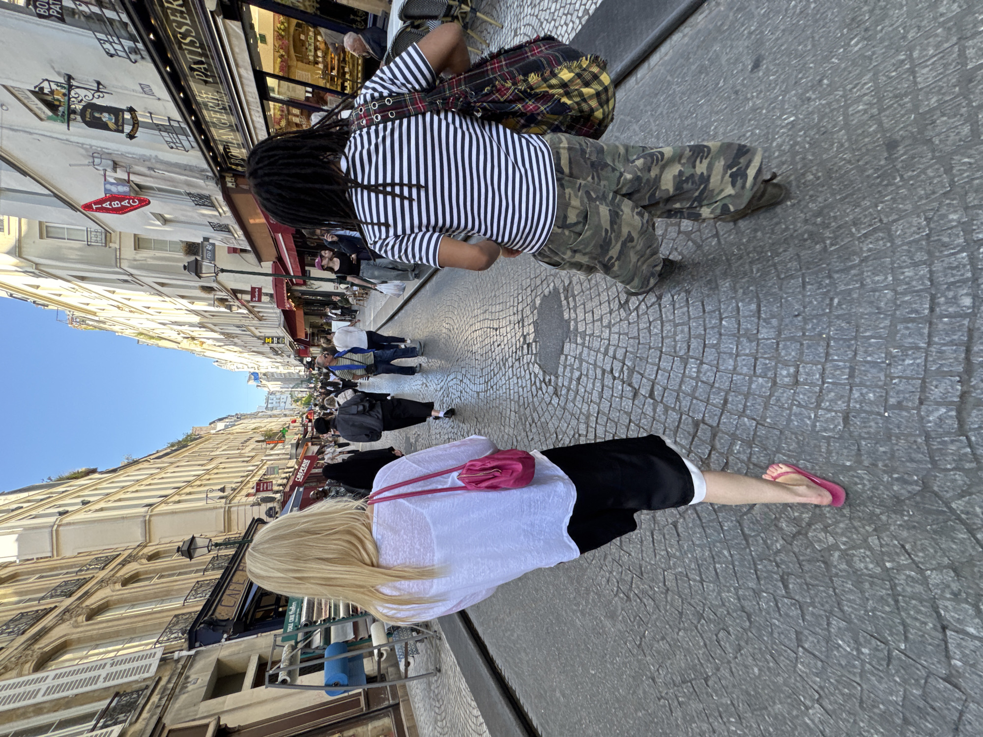 The image captures a vibrant street scene in Paris, France, under a clear blue sky, indicating bright daylight. The perspective looks down a narrow, cobblestone street flanked by multi-story buildings with classic Parisian architecture, including balconies and shopfronts.
In the immediate foreground, a person with long blonde hair is seen from behind, walking away from the camera. They wear a white top, black shorts or a skirt, and pink flip-flops, carrying a small pink purse over their shoulder.
Just ahead, another individual with dark dreadlocks is also walking away. They are dressed in a black and white striped t-shirt, camouflage pants, and a plaid backpack featuring yellow, red, and black tartan patterns.
Further down the street, several other pedestrians are visible, some walking, others possibly seated at outdoor cafes or browsing shops. The overall activity suggests a typical daytime bustle of people moving along the street.
Notable details include architectural elements like ornate ironwork on windows and balconies, as well as various shop awnings. Visible text on signs includes "BOULANGERIE PATISSERIE" on a prominent black sign, "TABAC" on a red oval sign, and "CAFE" on another storefront, all contributing to the French street ambiance. A blue rolled mat is also seen vertically against a building on the left.