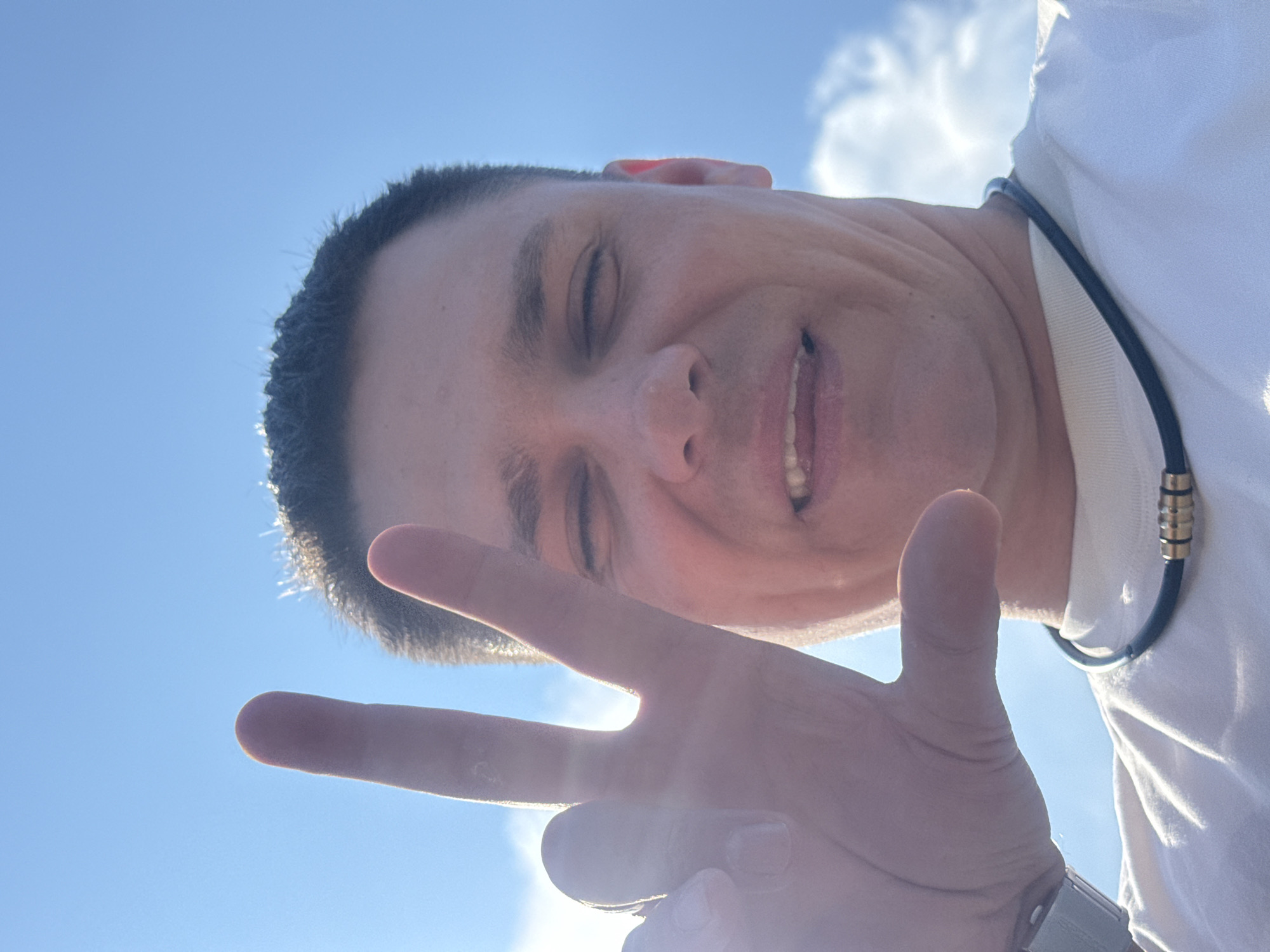 A man is taking a selfie outdoors, with a blue sky and some clouds in the background. He is wearing a white t-shirt and a black necklace with gold accents. His right hand is raised in front of the camera, with his index and middle fingers extended in a peace sign. His eyes are closed, and he has a slight smile on his face. The image is taken from a low angle, looking up at the man.
It is impossible to determine the specific location within the city of Vladivostok, Russia, based on the provided image alone.
