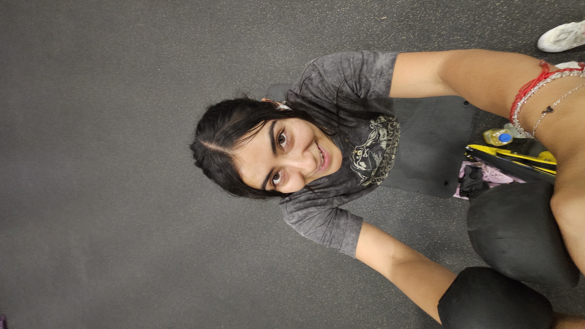 The image captures a high-angle shot of a young woman smiling brightly, likely taking a selfie or being photographed from above. She has dark hair, parted down the middle and pulled back, and is looking directly into the camera with a wide, friendly smile that shows her teeth. Her dark eyes appear cheerful.
She is wearing a dark grey, short-sleeved t-shirt with a faded, patterned graphic design on the chest, possibly featuring a skull or similar motif. On her right wrist, she sports multiple bracelets: a red string, a silver-colored beaded or chain bracelet, and another silver chain with a small dark charm. A white earbud is visible in her left ear. She appears to be wearing dark leggings or shorts, and a black exercise or yoga mat is beneath her.
The setting appears to be an indoor gym or fitness studio, indicated by the dark grey, textured rubber flooring. Various gym accessories are partially visible around her: a bright yellow object, possibly a water bottle with a blue cap, a pink and black item which could be a small bag or clothing, and part of a white and grey patterned shoe. Dark, indistinct objects, possibly weights, are near her legs. The lighting is bright and even, typical of an indoor facility.
The woman's pose and smile suggest she is happy and relaxed, likely taking a break during or after a workout. There is no legible text visible in the image. The location context provided is Puebla, Mexico.