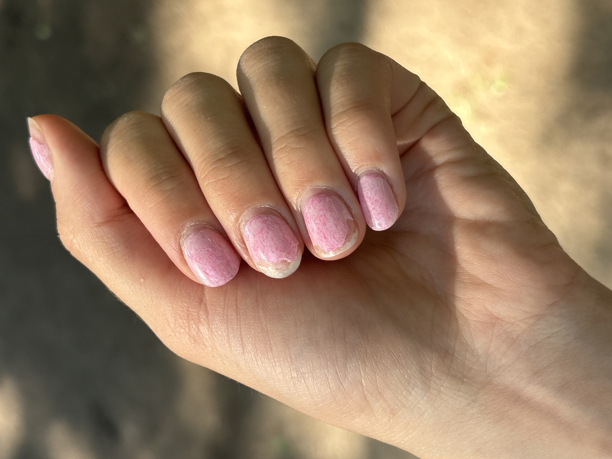 A fair-skinned human hand with chipped pink nail polish is shown in an outdoor setting. The hand is positioned palm-upward, fingers slightly bent, with light and shadow playing across the skin. The background is blurred, but appears to be a natural environment with shades of brown and green, possibly a park or wooded area. There is no evidence that this photo was taken in Perm, Russia.