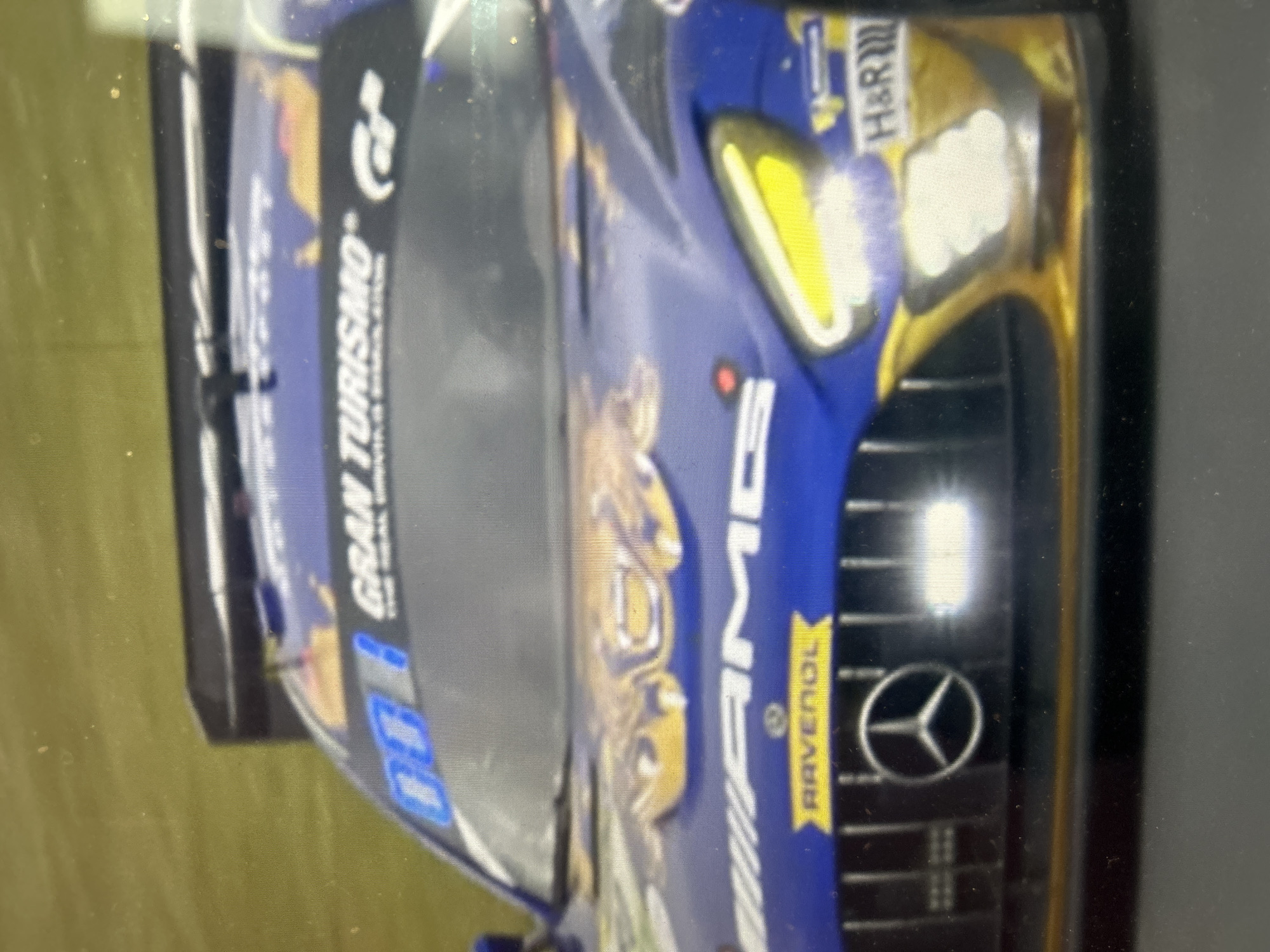 The image shows a blue and gold Mercedes-AMG GT3 race car. It features branding for Gran Turismo, AMG, Ravenol, and H&R. The location is not discernible from the image.