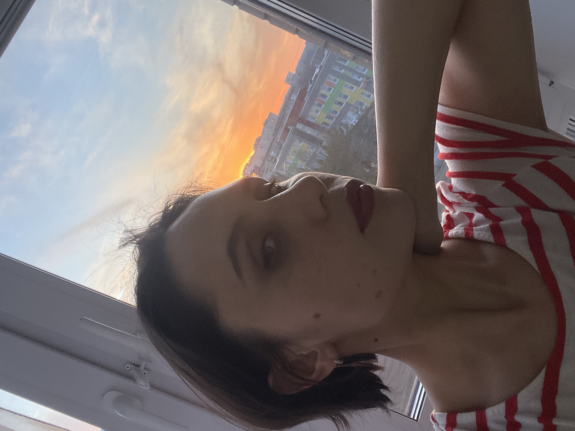 A woman with dark hair and red lipstick is lying on her side, looking toward a window. The window reveals a sunset sky with hues of orange and blue, and a building in the distance. The building has a colorful facade with sections of green, yellow, blue, and red. The woman is wearing a red and white striped shirt. The location is Izhevsk, Russia.