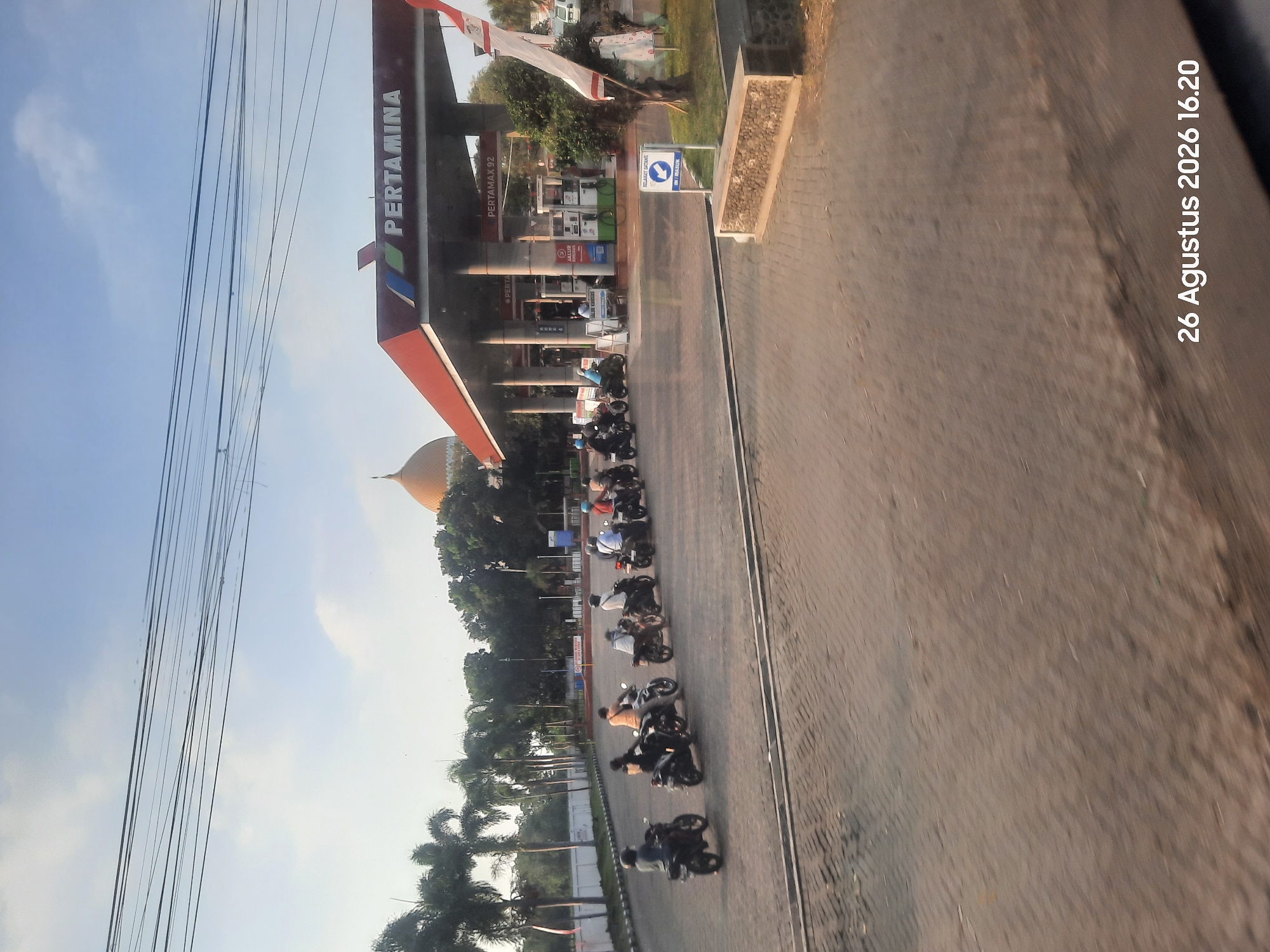 The image depicts a Pertamina gas station in Klaseman, Indonesia, on August 26, 2026, at 16:20. A line of motorcycles, each with a rider, is queuing at the station. Palm trees and other foliage surround the area, with a large golden dome visible in the background, likely a significant landmark. The sky is clear with scattered clouds, and power lines stretch across the upper portion of the frame. The ground is paved with interlocking stones. A sign indicating "SELAMAT DATANG" (Welcome) and "MASUK" (Entrance) with an arrow is visible. The gas station features multiple pumps and a canopy with the Pertamina logo. A white and red flag is also present. The lighting suggests it is late afternoon.