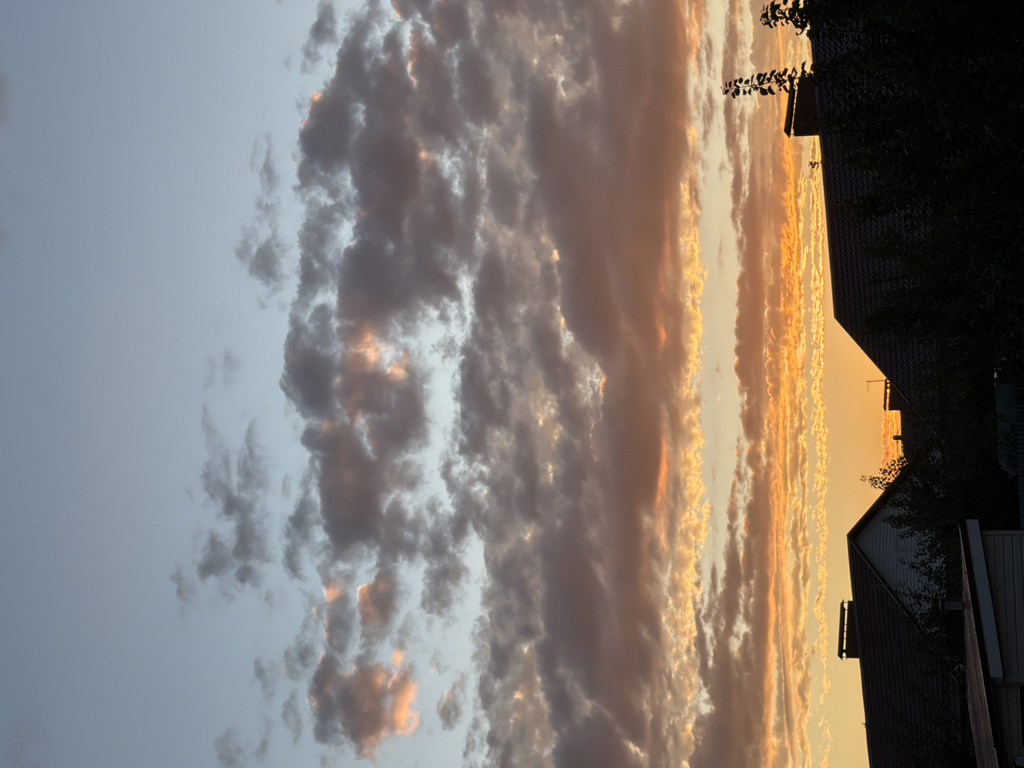 The image captures a sunset over a residential area in Uzyukovo, Russia. The sky is filled with dramatic clouds, ranging in color from pale blue at the top to vibrant oranges and yellows near the horizon. The clouds are illuminated by the setting sun, casting warm hues across the scene. In the foreground, the dark silhouettes of rooftops and trees are visible, indicating houses and vegetation. One house in the center has a prominent chimney. The overall atmosphere is serene and visually striking due to the intense colors of the sunset.