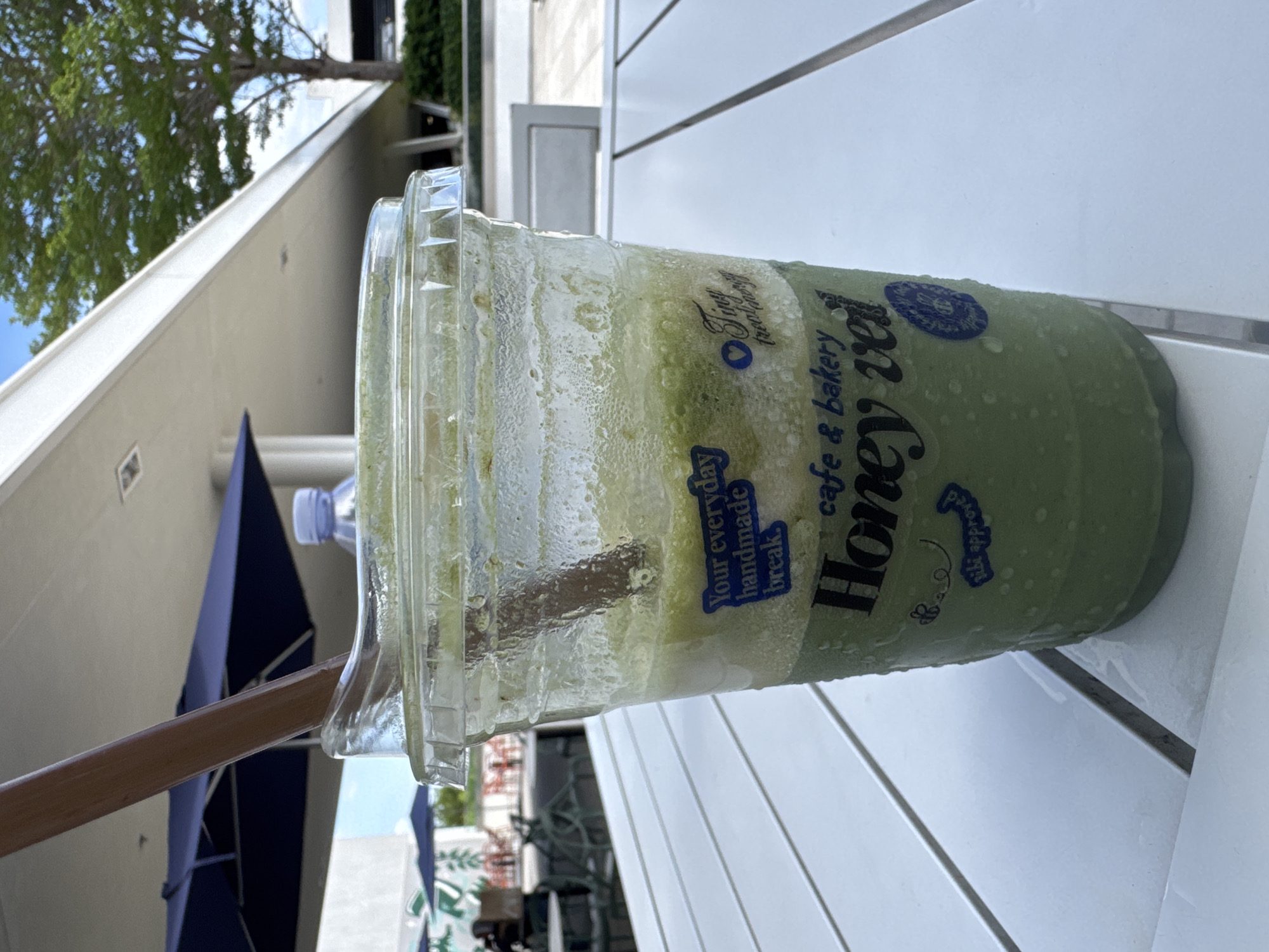 An outdoor scene in Hallandale Beach, Florida, United States of America, shows a close-up of a green, blended beverage in a clear plastic cup with a lid and a wooden straw. The cup has blue text on it that reads "Your everyday handmade break," "Tiny treat shop," "cafe & bakery," and "Honey Veil." There is also a small bee illustration with the text "uki approved." The cup is condensation-covered and sits on a white slatted table. In the background, there are blue umbrellas, a tree, and a modern building. The lighting suggests it is daytime.